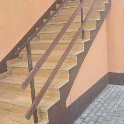 Bogdan Bróg - Zewnętrzne schody wyłożone płytkami imitującymi drewno, z brązową balustradą i cokołem. Ściana w odcieniach brzoskwini, obok kostka brukowa. Ujęcie z dołu, perspektywa.