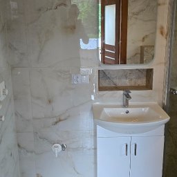 Bogdan Bróg - Wykończona łazienka z umywalką na szafce, lustrem w zaokrąglonej ramie i marmurowymi płytkami na ścianach i podłodze. Widoczne gniazdka elektryczne.