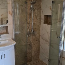 Bogdan Bróg - Wykończona łazienka z kabiną prysznicową typu walk-in, marmurowymi płytkami, białą umywalką z szafką i ozdobną lampą sufitową w kształcie kwiatu.