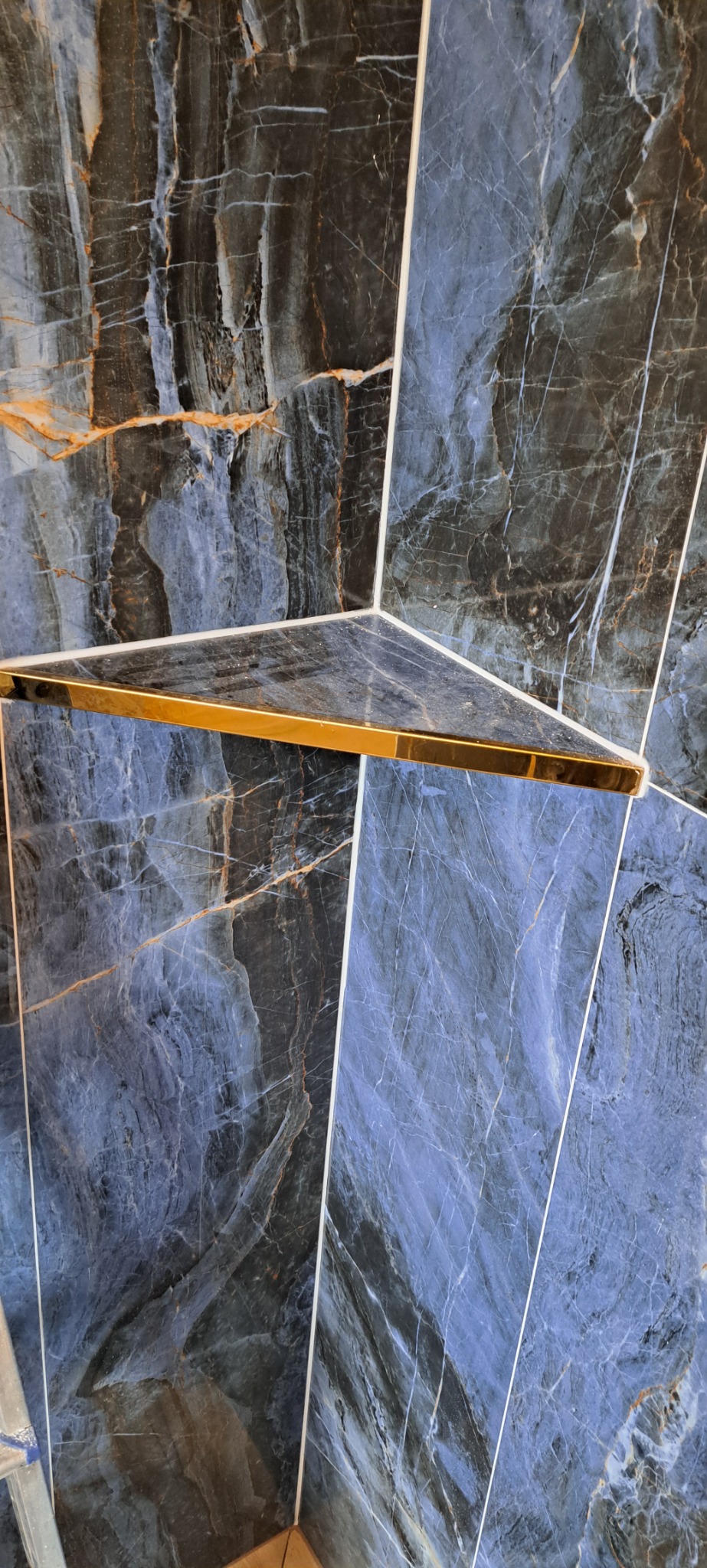 Niebiesko-czarne płytki imitujące marmur z wyraźnymi żyłami, ułożone pod kątem, z półką narożną z akcentem w kolorze złotym.