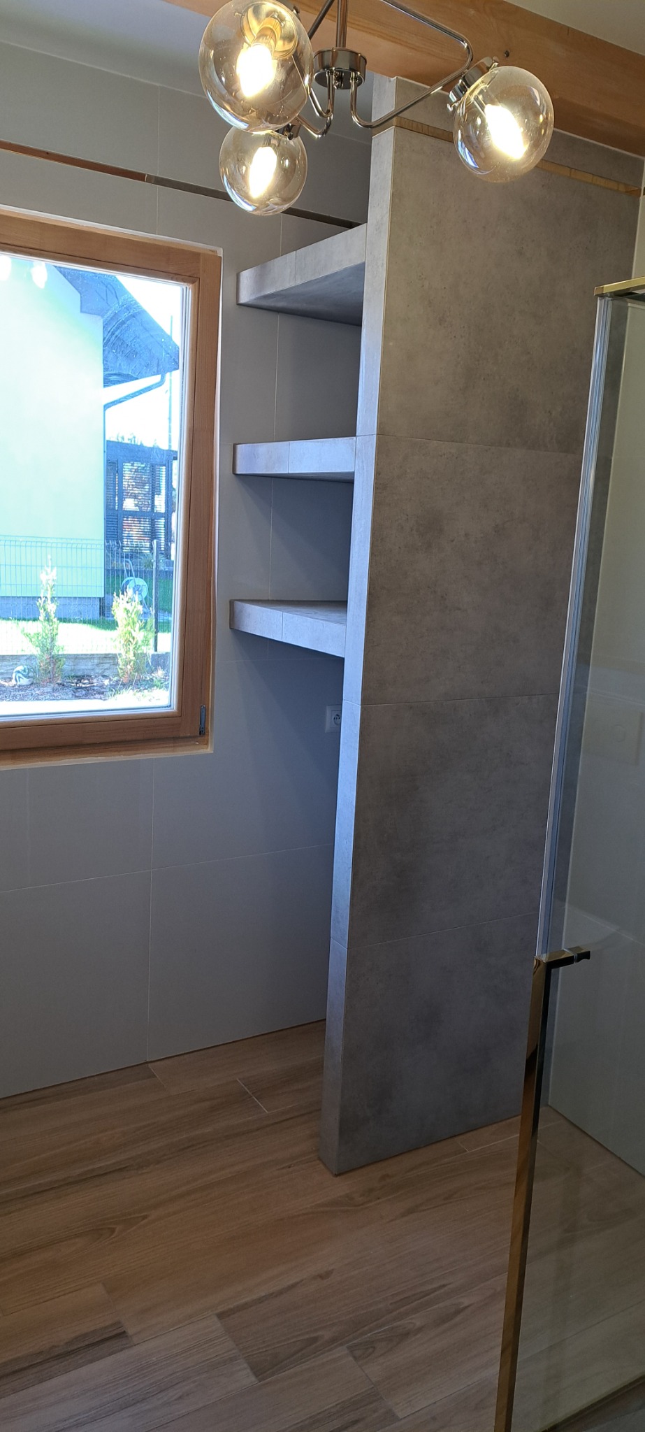 Nowoczesna łazienka z drewnopodobną podłogą, szarymi płytkami na ścianach i wnęką z półkami z płytek, oświetlona lampą sufitową z kulistymi kloszami. Widok przez okno na zewnątrz.