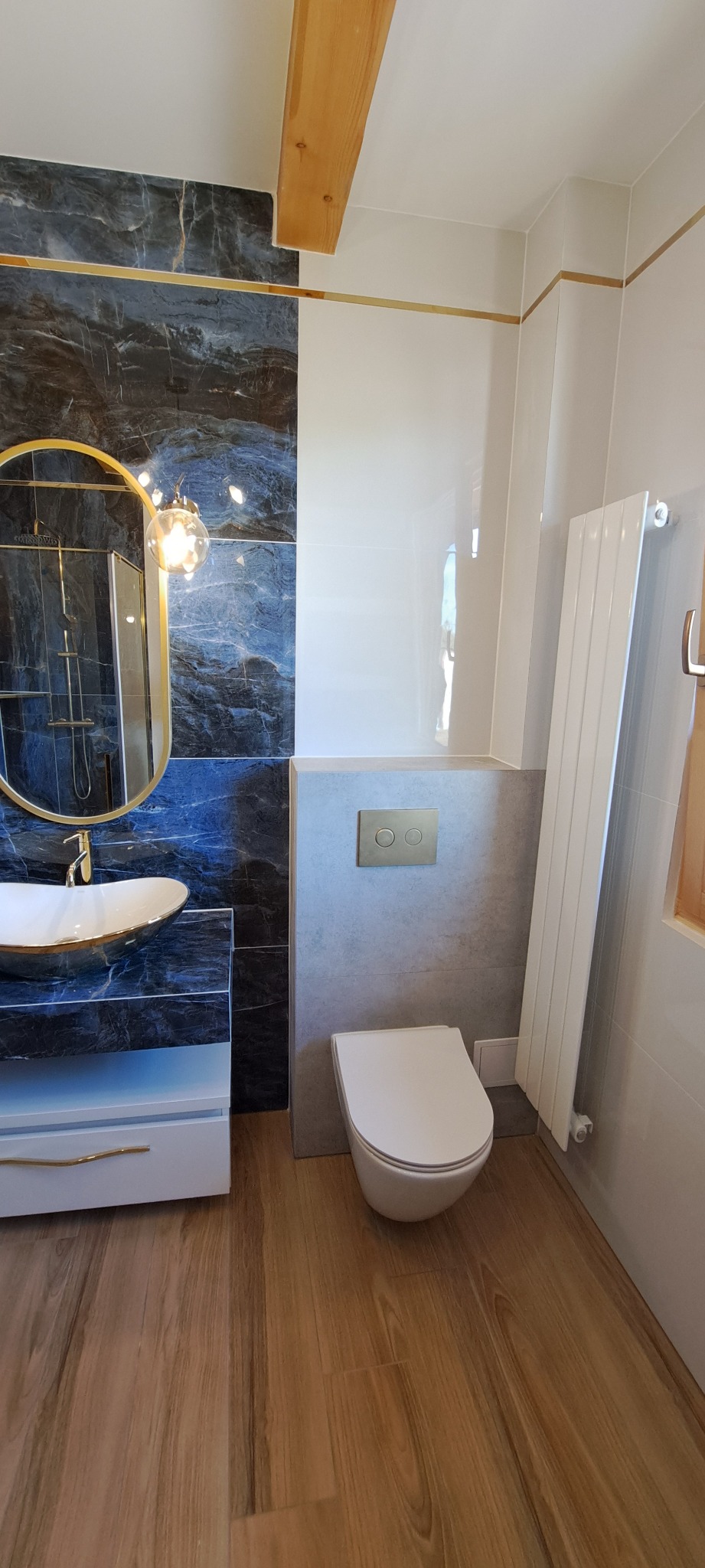 Nowoczesna łazienka z umywalką nablatową w kolorze białym ze złotymi akcentami, okrągłym lustrem w złotej ramie, toaletą wiszącą i grzejnikiem panelowym, wykończona płytkami imitującymi marmur...