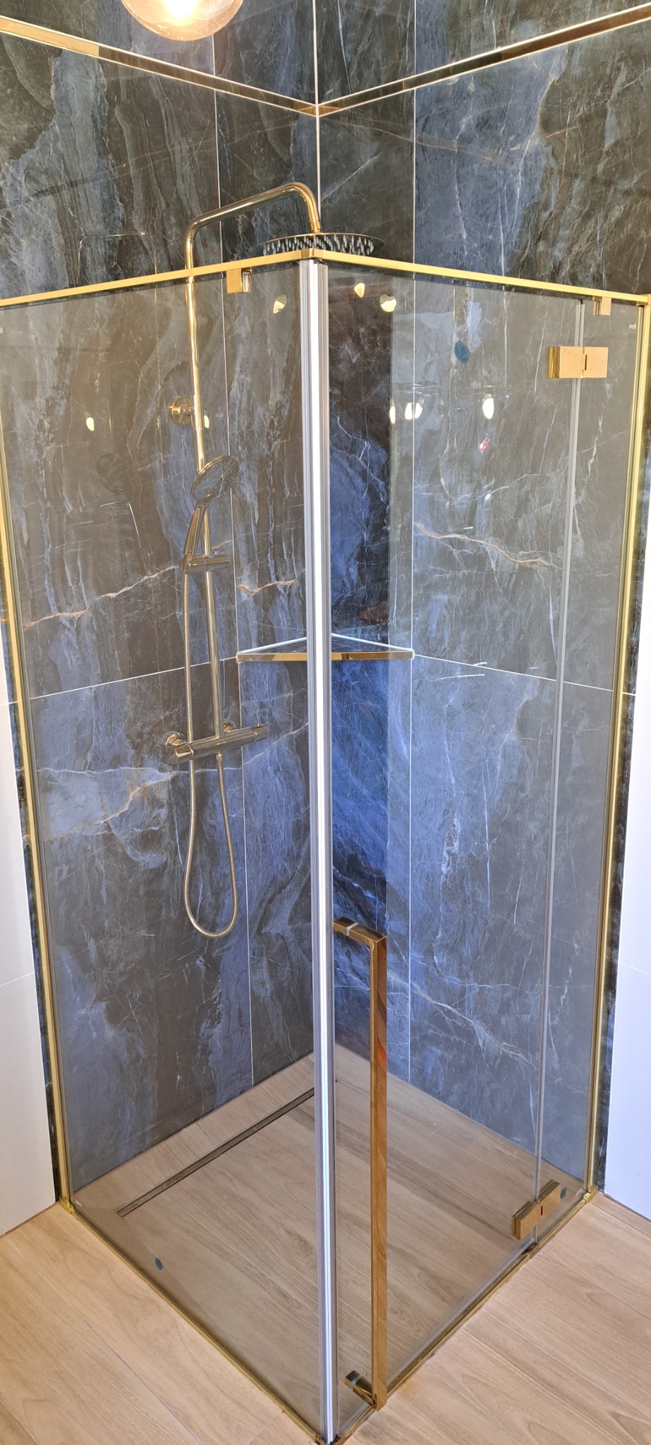 Nowoczesna kabina prysznicowa z złotymi detalami, marmurowe płytki w odcieniach szarości, drewnopodobna podłoga, oświetlenie sufitowe.