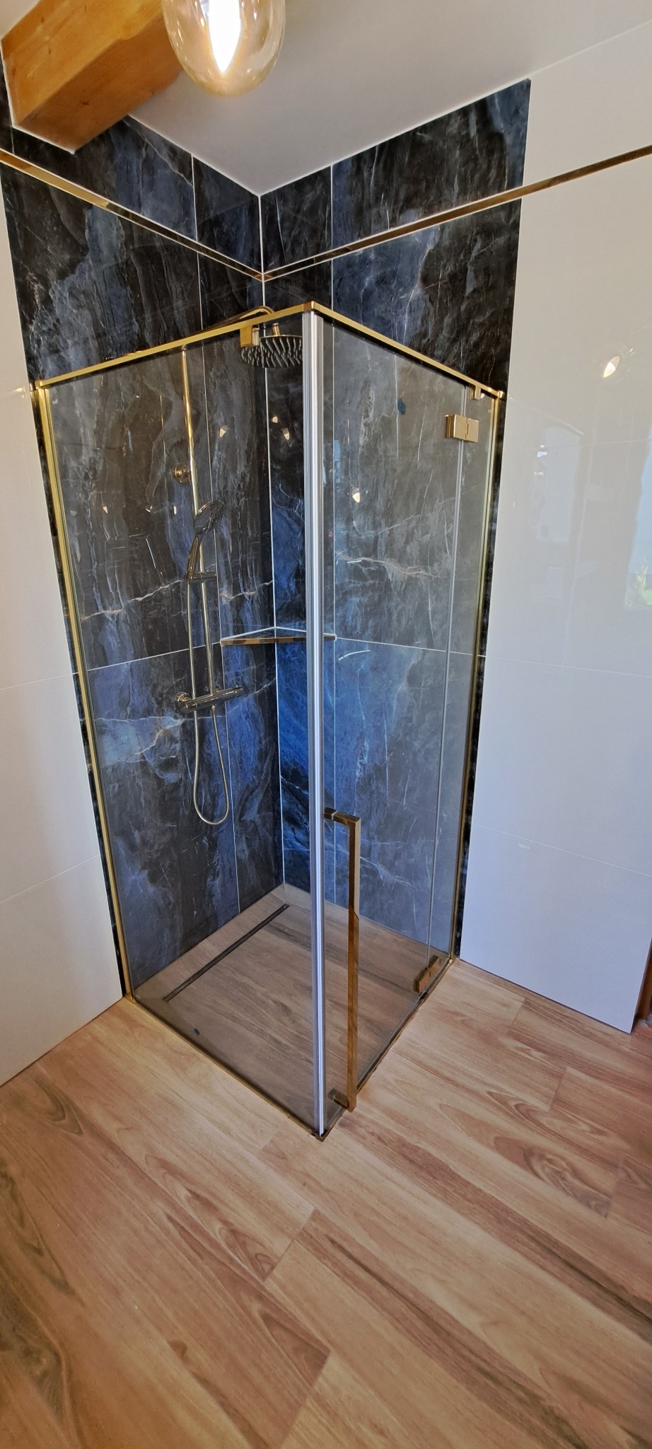 Nowoczesna kabina prysznicowa z czarnymi marmurkowymi płytkami i złotymi detalami, drewnopodobna podłoga, oświetlenie górne podkreśla luksusowy charakter.