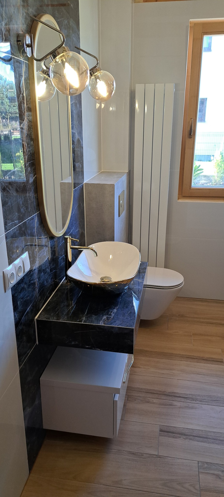 Nowoczesna łazienka z umywalką nablatową w kolorze białym z złotymi akcentami, czarnym blatem imitującym marmur, owalnym lustrem z oświetleniem i drewnopodobną podłogą.