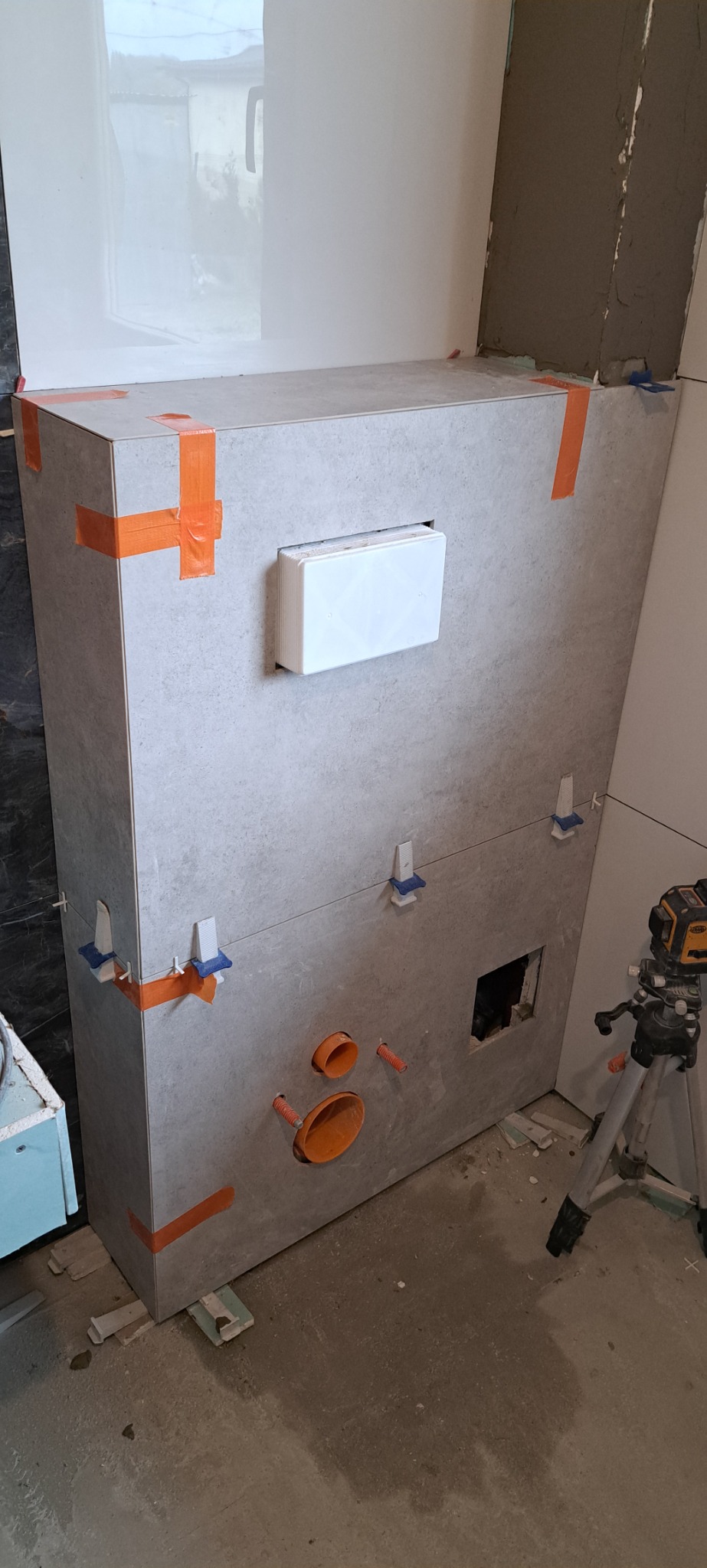 Szara obudowa podtynkowego WC w trakcie montażu płytek, z widocznymi otworami na rury, poziomica laserowa na trójnogu, pomarańczowa taśma klejąca i klipsy do płytek.