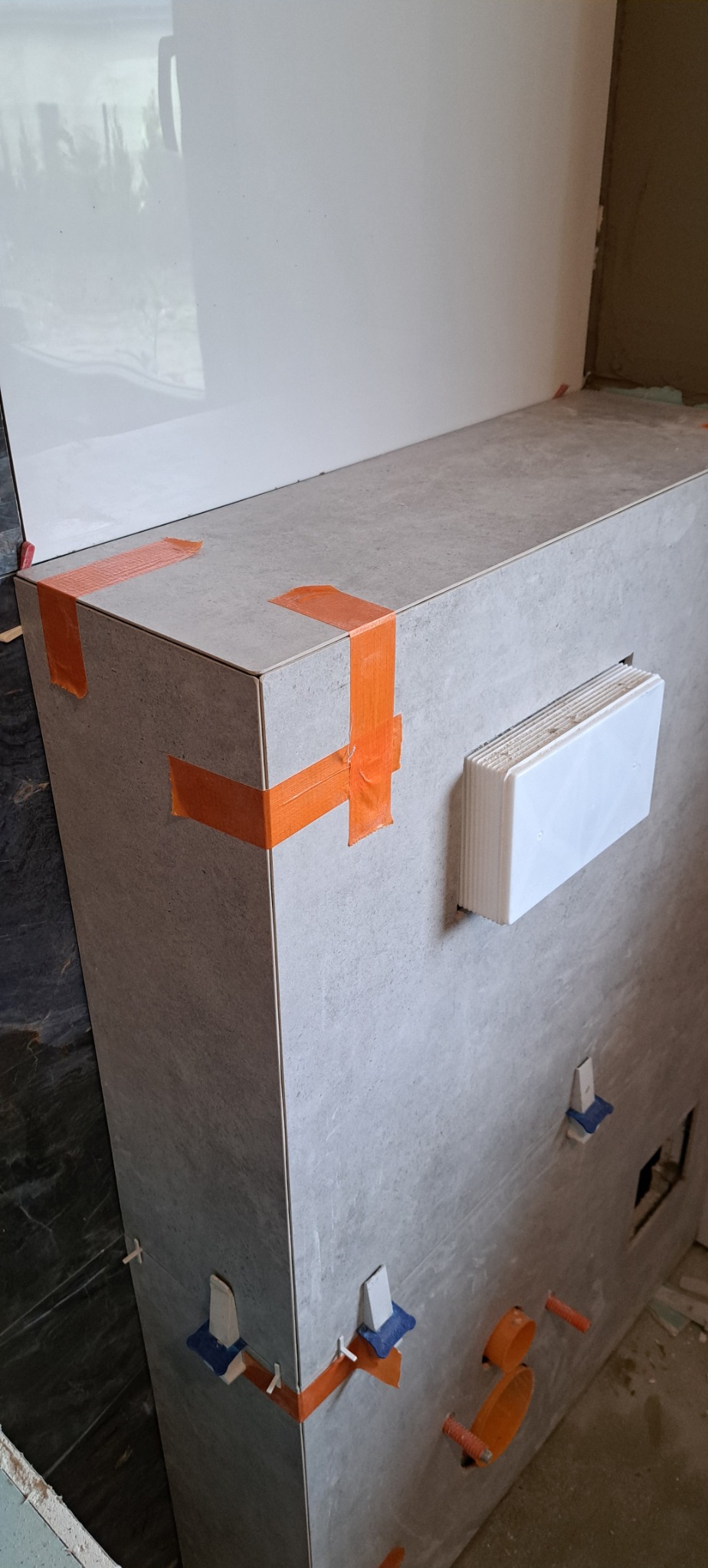 Szara zabudowa z płytek w trakcie układania, zabezpieczona pomarańczową taśmą, widoczne krzyżyki dystansowe i otwory na instalacje.
