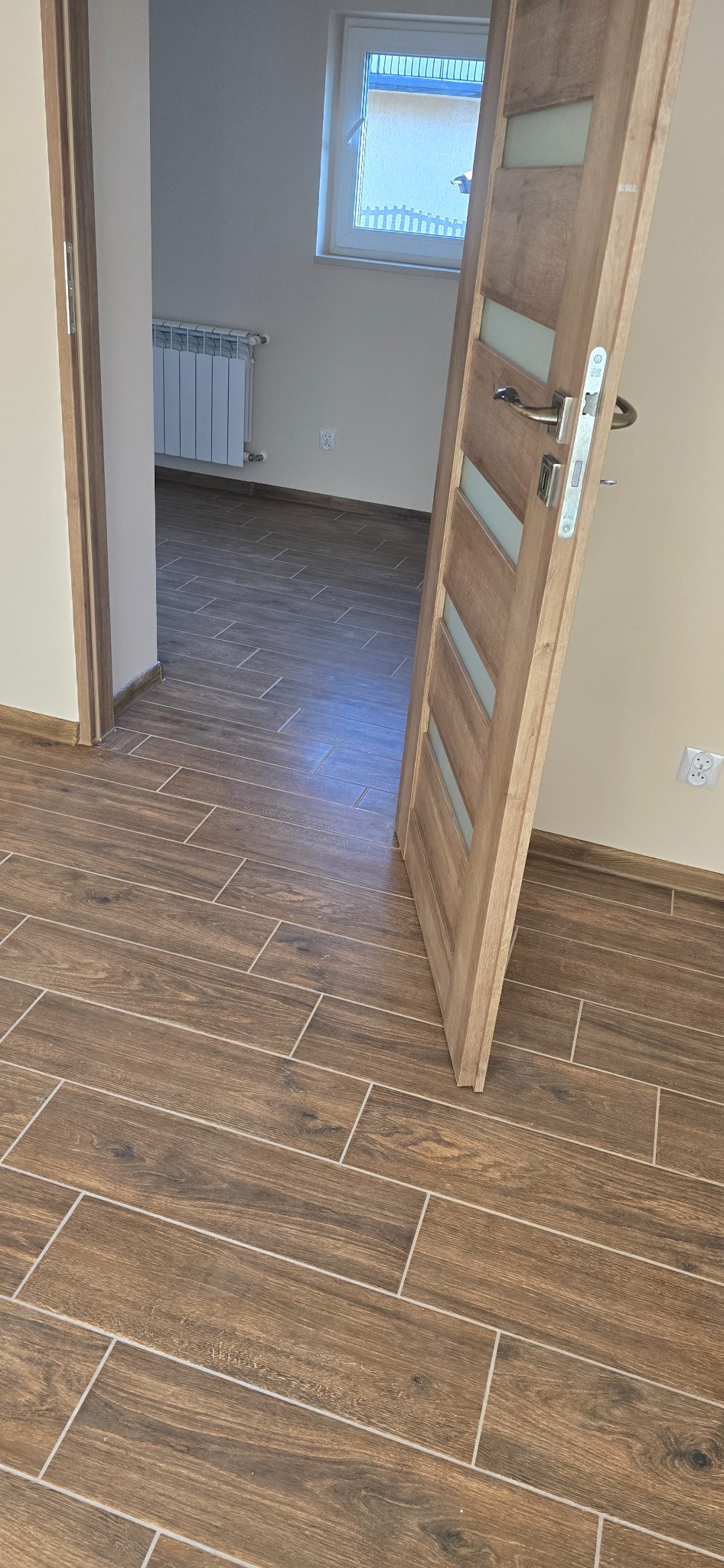 Ułożona podłoga z płytek imitujących drewno w nowym mieszkaniu, widoczny fragment białego grzejnika, białe ściany i drewniane drzwi z szybami z mlecznego szkła