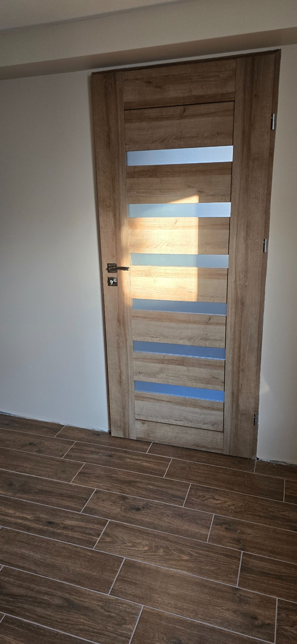 Zamontowane wewnętrzne drzwi z drewnianą okleiną i poziomymi, matowymi szybami w Miedzianej Górze, widoczne na tle brązowej podłogi z płytek imitujących drewno i świeżo pomalowanej ściany.