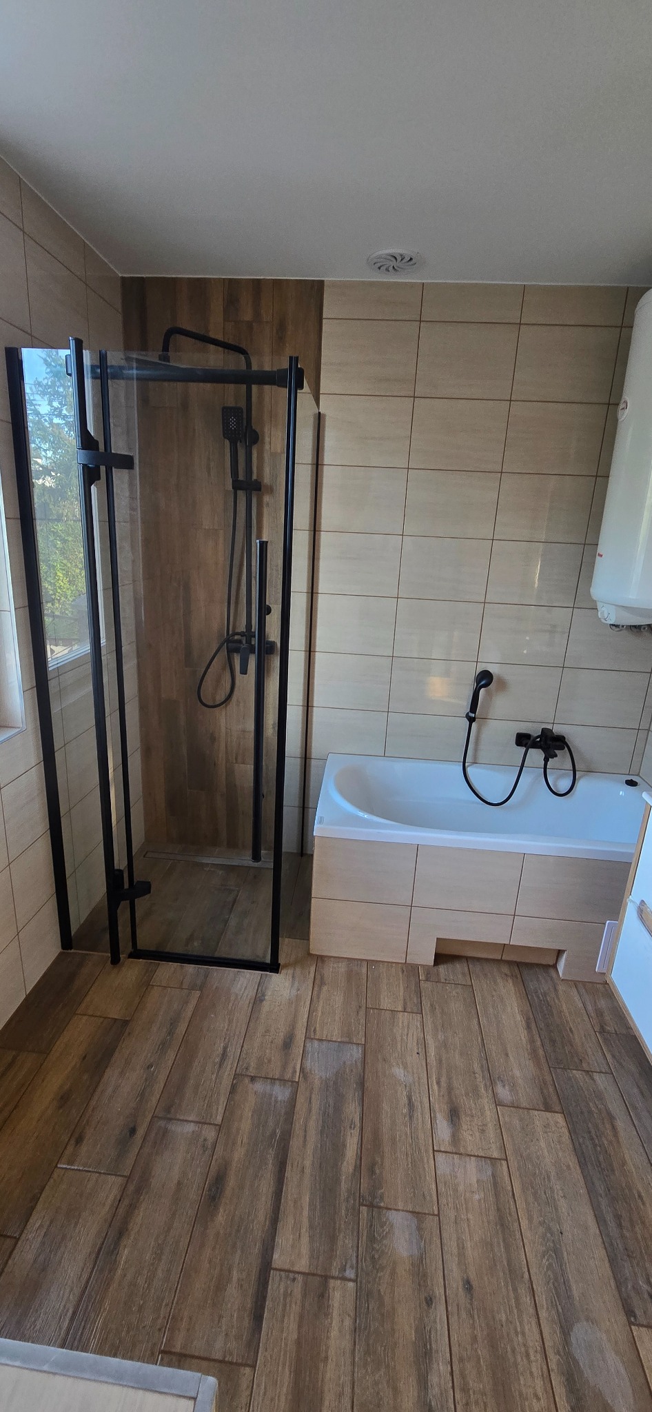 Nowoczesna łazienka z kabiną prysznicową typu walk-in z czarnymi profilami, wanną obudowaną płytkami i podłogą imitującą drewno.