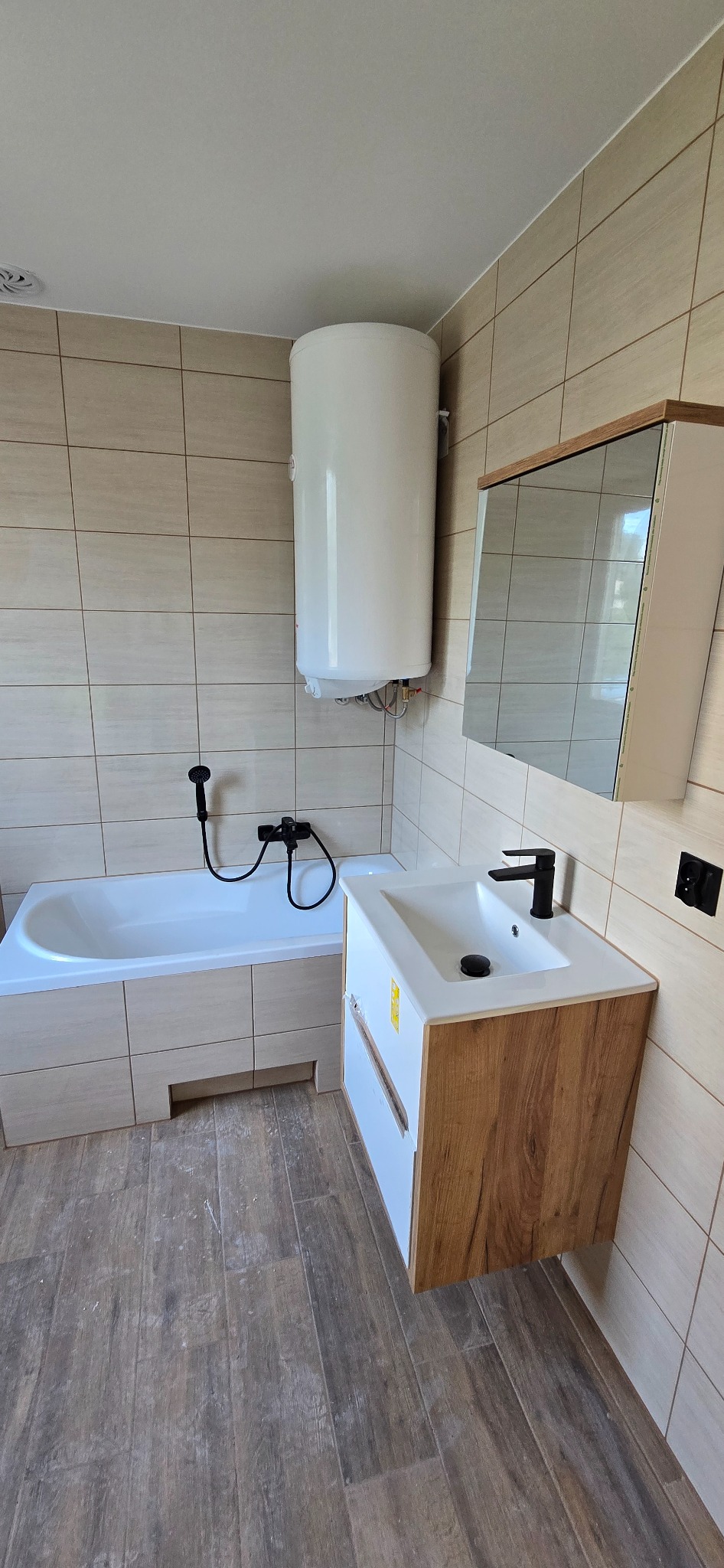 Jasna łazienka po remoncie: beżowe płytki, wanna z obudową, umywalka z szafką z drewnianym wzorem, bojler. Minimalistyczny styl, czarna armatura.