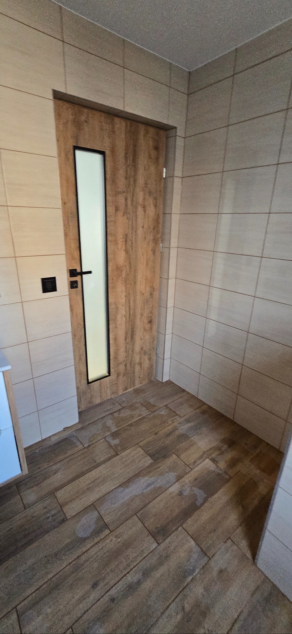 Wykończona łazienka z drewnopodobnymi płytkami na podłodze i ścianach. Widoczne drzwi z szybą i czarną klamką oraz fragment białej szafki.