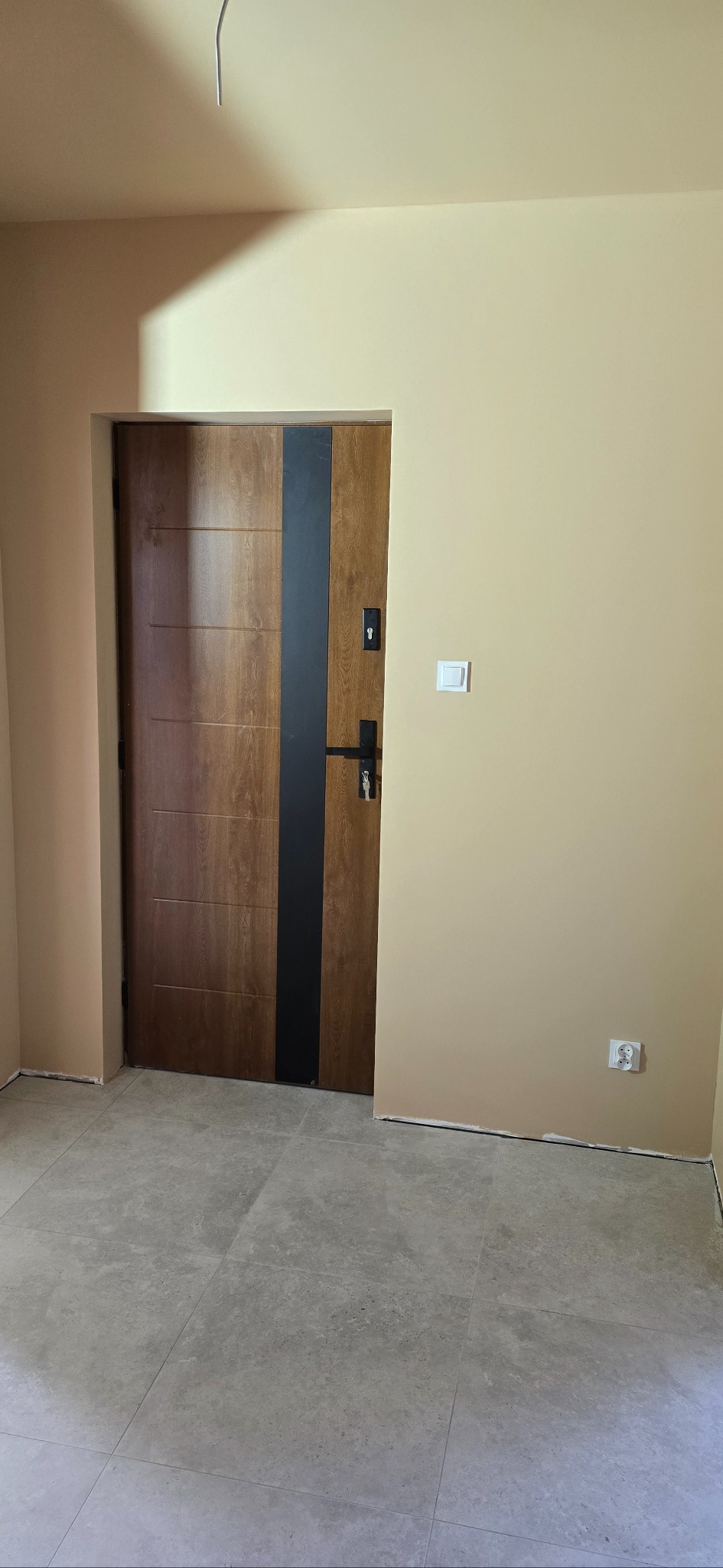 Nowoczesne drzwi wejściowe z drewnopodobnym wykończeniem i czarnym akcentem, zamontowane w jasnym wnętrzu z szarymi płytkami podłogowymi. Widoczny włącznik i gniazdko.