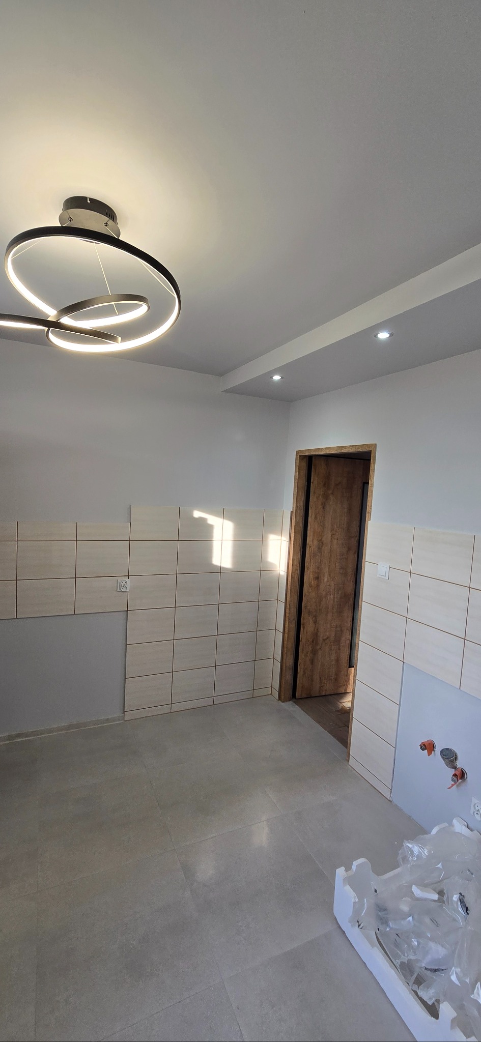 Wnętrze w trakcie wykańczania: ułożone płytki na ścianie do połowy wysokości, szare płytki na podłodze, nowoczesna lampa sufitowa i drewniane drzwi.