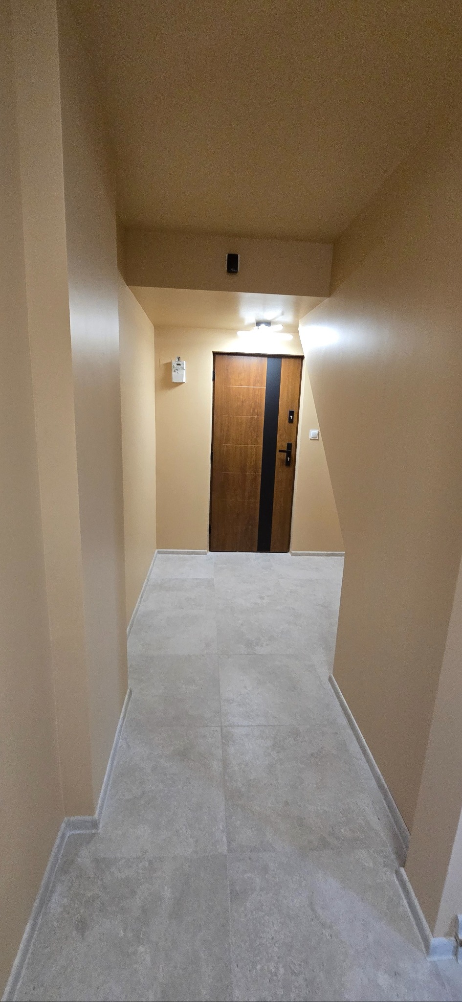 Beżowy korytarz po remoncie z szarą podłogą z płytek i drewnianymi drzwiami z czarnym paskiem. Widok z perspektywy.
