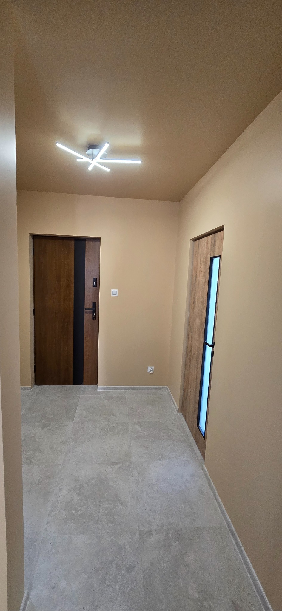 Beżowy przedpokój z dwiema drewnianymi drzwiami, szarą podłogą z płytek i nowoczesnym oświetleniem sufitowym.