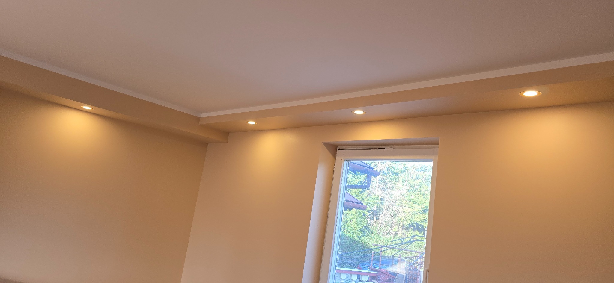 Jasne wnętrze po remoncie: beżowe ściany, biały sufit z oświetleniem punktowym i okno z widokiem na zieleń, wykończone listwy przysufitowe.