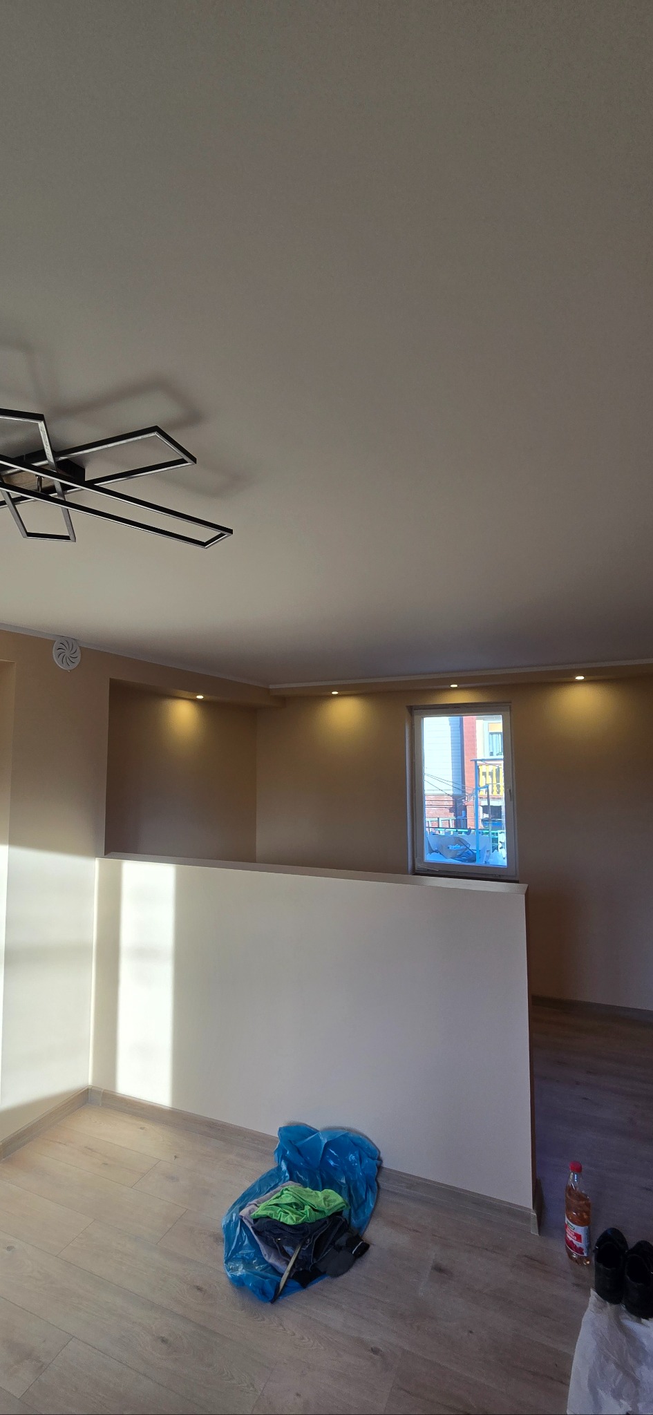 Jasne wnętrze po remoncie: kremowe ściany, panele podłogowe, nowoczesna lampa sufitowa. Widoczne ślady po pracach wykończeniowych, okno z widokiem na budynek.