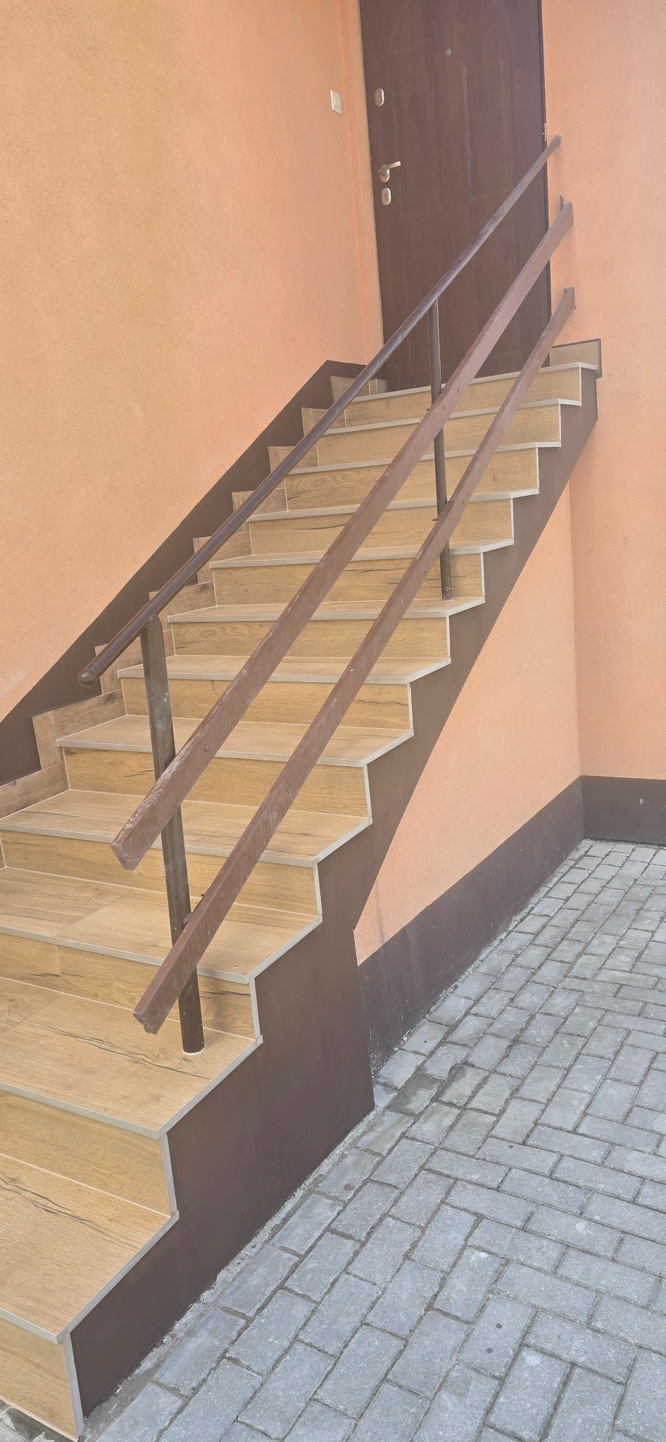 Zewnętrzne schody wyłożone płytkami imitującymi drewno, z brązową balustradą i cokołem. Ściana w odcieniach brzoskwini, obok kostka brukowa. Ujęcie z dołu, perspektywa.