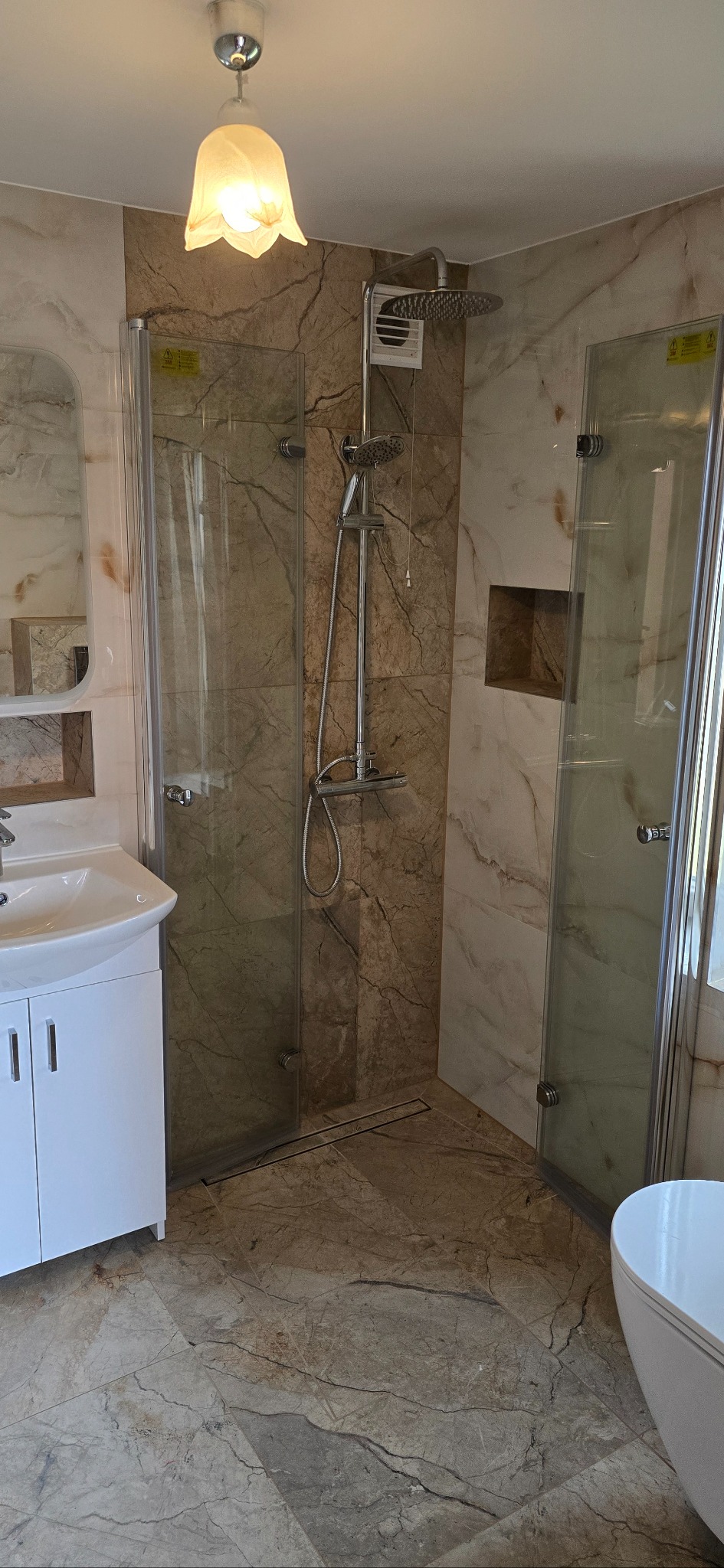 Wykończona łazienka z kabiną prysznicową typu walk-in, marmurowymi płytkami, białą umywalką z szafką i ozdobną lampą sufitową w kształcie kwiatu.