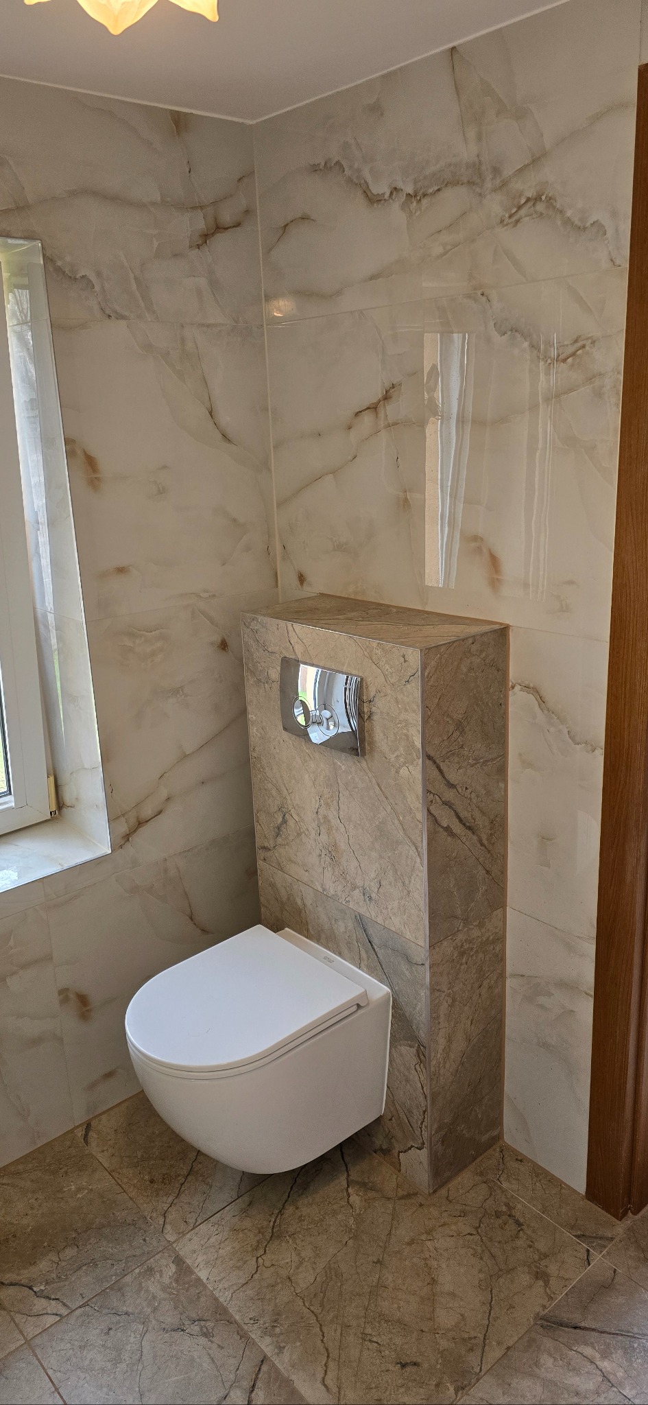 Wykończona łazienka z podwieszaną toaletą w obudowie z płytek imitujących marmur. Płytki ścienne i podłogowe w podobnym stylu. Widoczne okno i fragment drewnianej ościeżnicy.