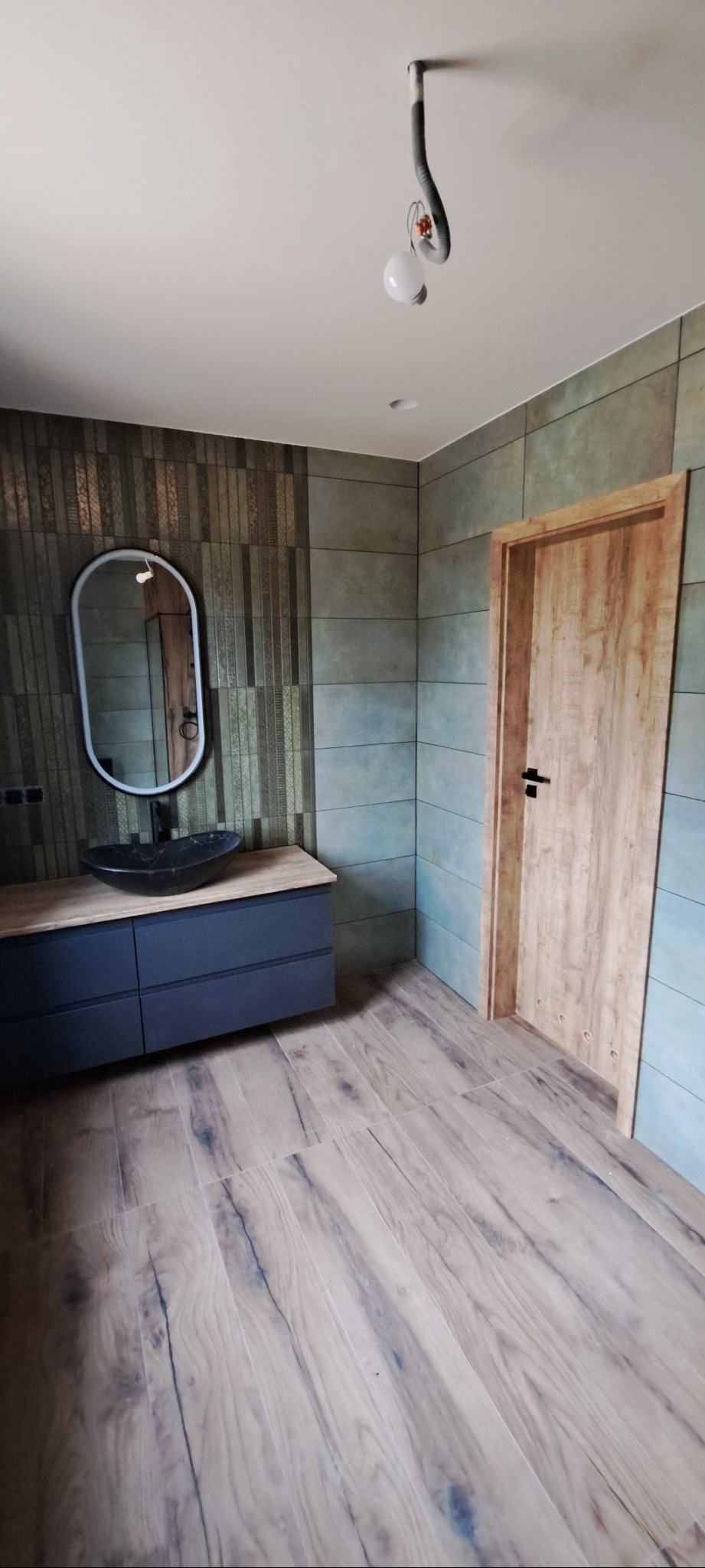 Nowoczesna łazienka z drewnianą podłogą i drzwiami, umywalką na szafce i owalnym lustrem. Ściany wyłożone płytkami w odcieniach szarości i zieleni.