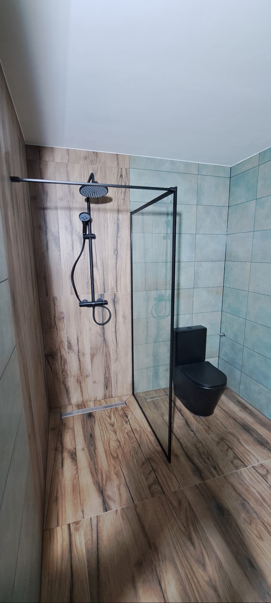 Nowoczesna łazienka z kabiną typu walk-in, czarną armaturą i toaletą. Płytki imitujące drewno na podłodze i ścianie, akcent turkusowych płytek w tle.