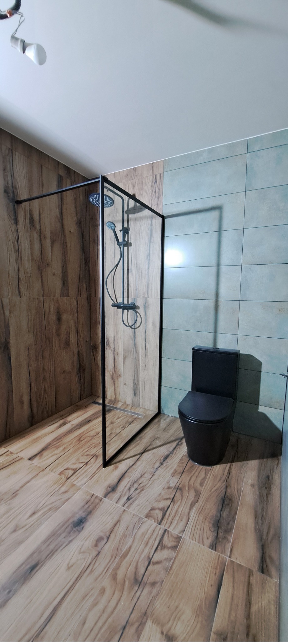 Nowoczesna łazienka z kabiną prysznicową typu walk-in z czarną ramą i czarną toaletą. Płytki imitujące drewno na podłodze i ścianie. Akcent turkusowych płytek.