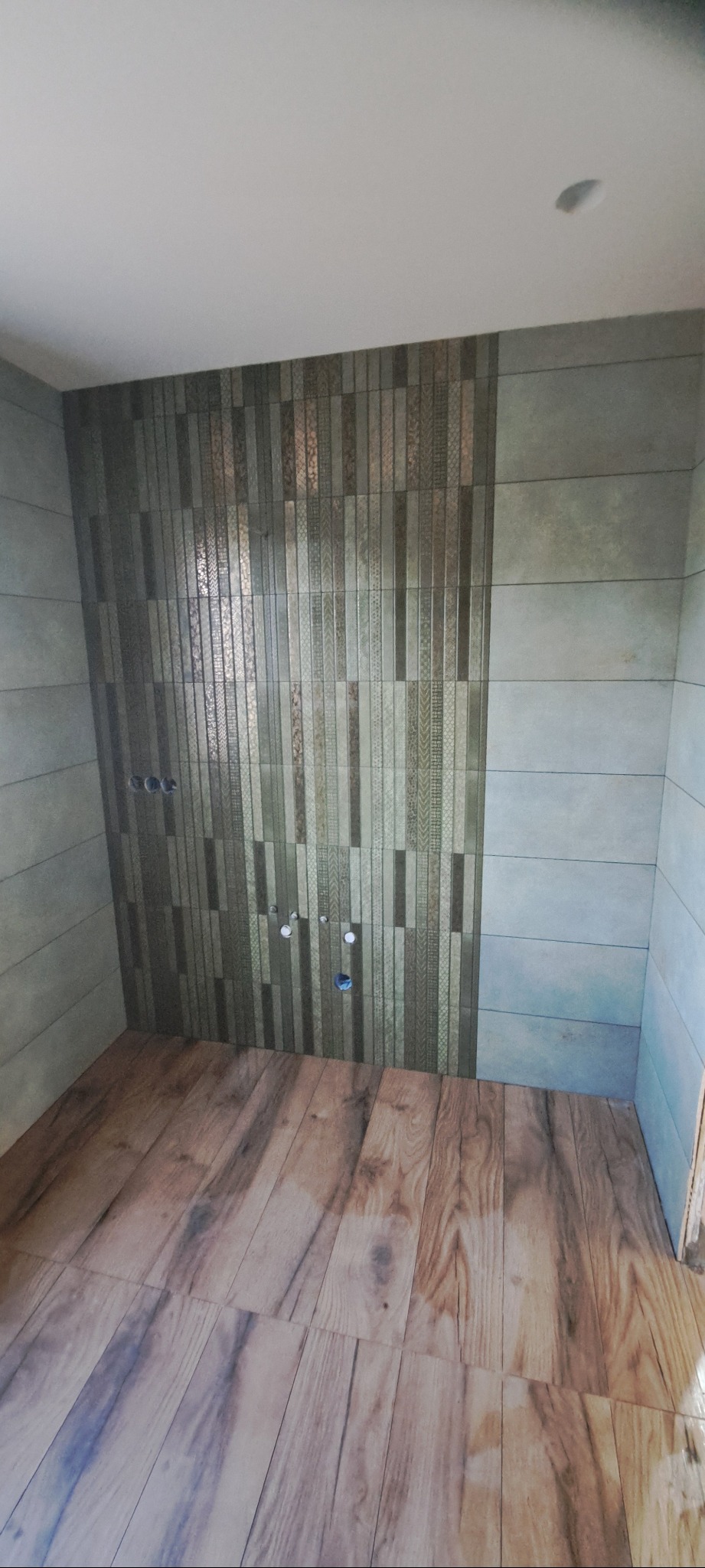 Wykończona łazienka z drewnopodobną podłogą i mozaiką na ścianie. Widoczne otwory na armaturę. Ukończony etap prac wykończeniowych.