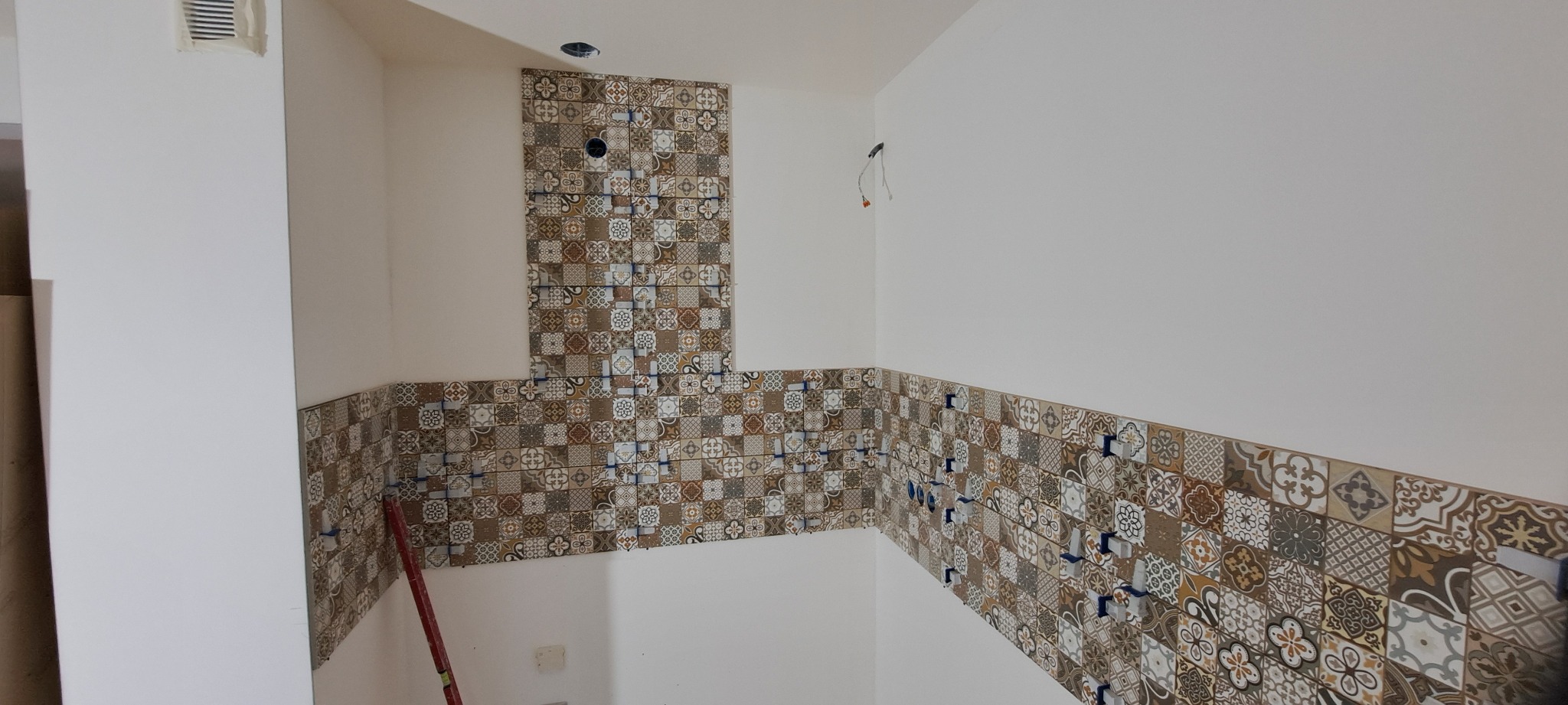 Układanie dekoracyjnych płytek patchwork na ścianie w łazience. Widoczne krzyżyki dystansowe. Częściowo wykończona ściana z otworem na instalację.