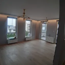 Jasne, puste wnętrze mieszkania po remoncie, z parkietem, grzejnikami pod oknami i nowoczesnymi lampami sufitowymi z kulistymi kloszami.