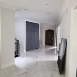 Wnętrze domu po remoncie: białe ściany, marmurowe podłogi, szara zabudowa szaf wnękowych, widoczny fragment schodów i czarna lodówka side-by-side, obraz oparty o ścianę.