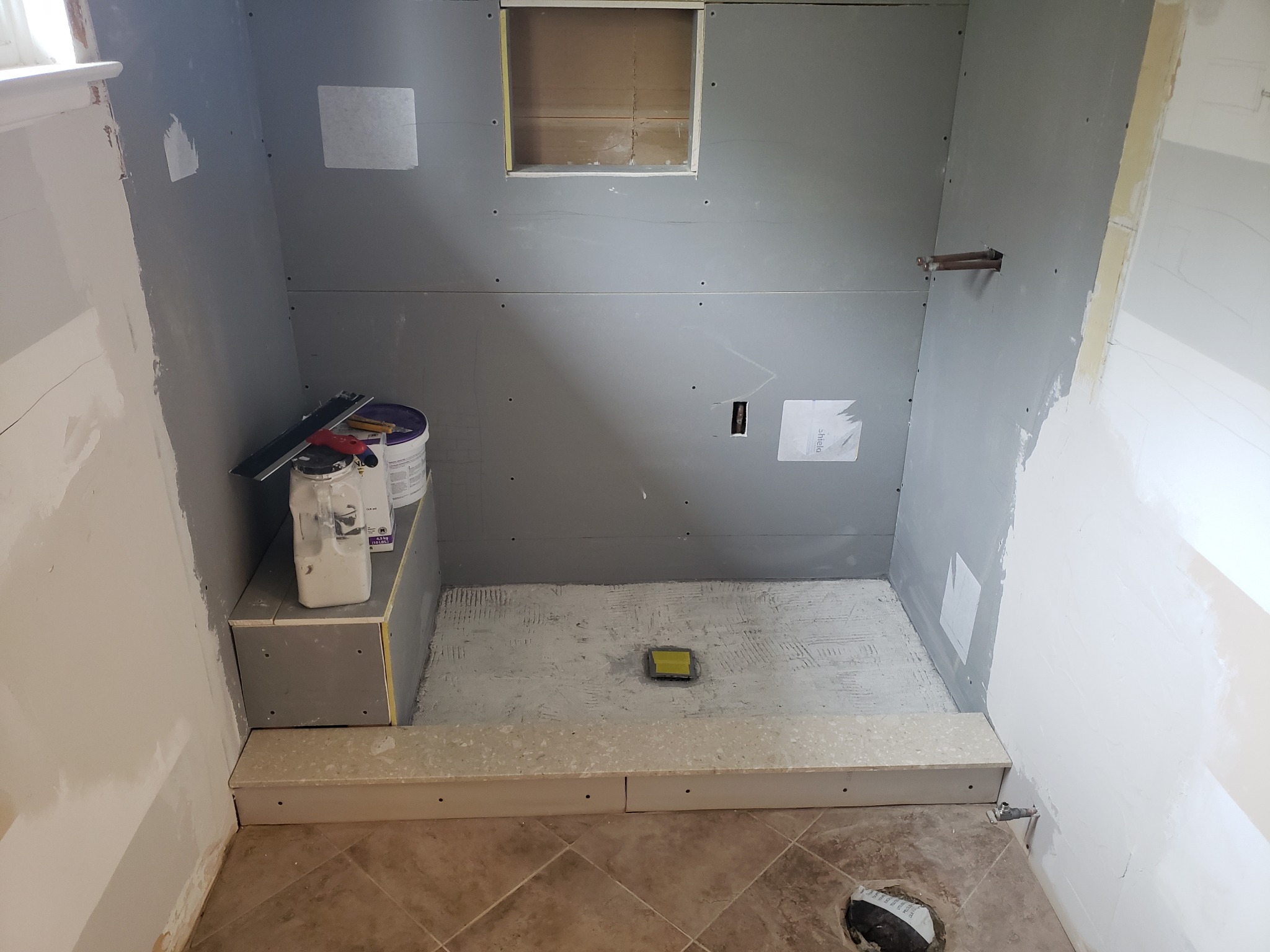 Wnętrze kabiny prysznicowej w trakcie budowy; widoczne płyty kartonowo-gipsowe na ścianach, przygotowane podłoże pod płytki na podłodze, odpływ liniowy zabezpieczony folią, oraz narzędzia...