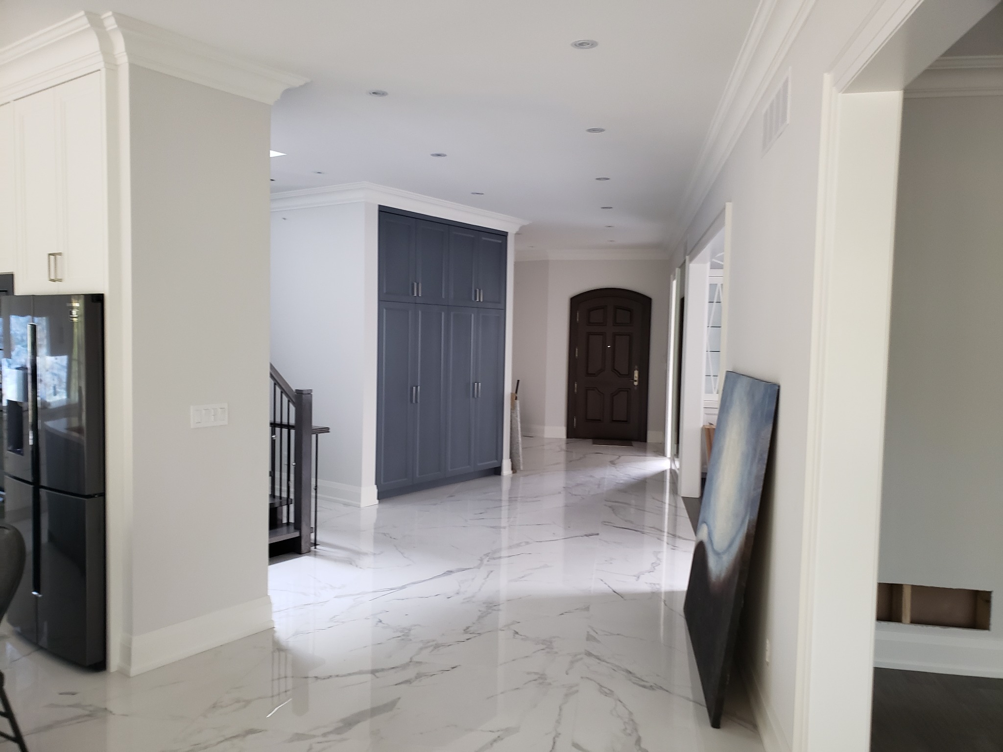 Wnętrze domu po remoncie: białe ściany, marmurowe podłogi, szara zabudowa szaf wnękowych, widoczny fragment schodów i czarna lodówka side-by-side, obraz oparty o ścianę.