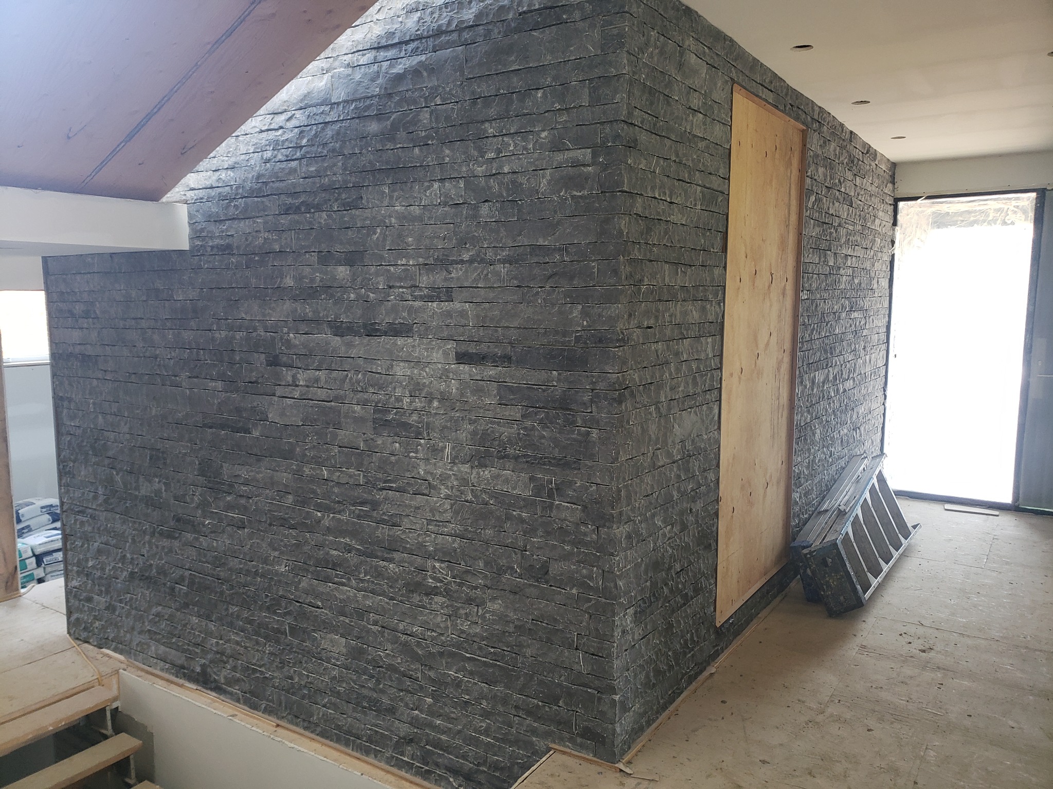 Ściana wewnętrzna wykończona czarnym kamieniem dekoracyjnym ułożonym w poziome pasy, widoczny fragment drewnianego sufitu i drzwi zabezpieczone płytą, drabina oparta o ścianę, w tle worki...