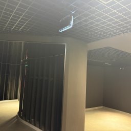 Gr Golden Renovations - Wnętrze w trakcie wykańczania: sufit rastrowy, ściany w ciemnym kolorze, widoczne elementy konstrukcyjne. Oświetlenie sufitowe. Minimalistyczny design w surowym stanie.