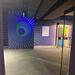 Gr Golden Renovations - Abstrakcyjna instalacja artystyczna: ściana z neonowym wzorem, gablota z ruchomą konstrukcją i betonowa podłoga w minimalistycznym wnętrzu.