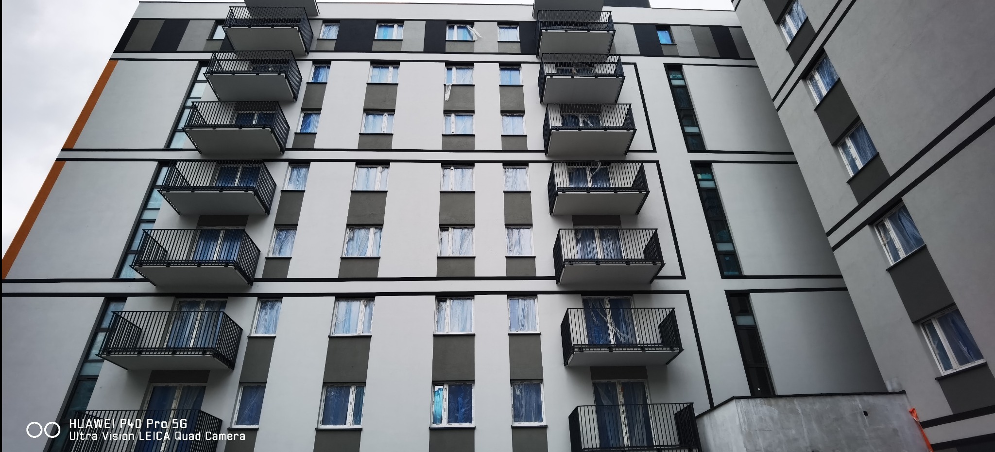 Elewacja nowego bloku z balkonami i oknami zabezpieczonymi folią. Widok z dołu, podkreślający geometrię budynku i etap wykończeniowy.