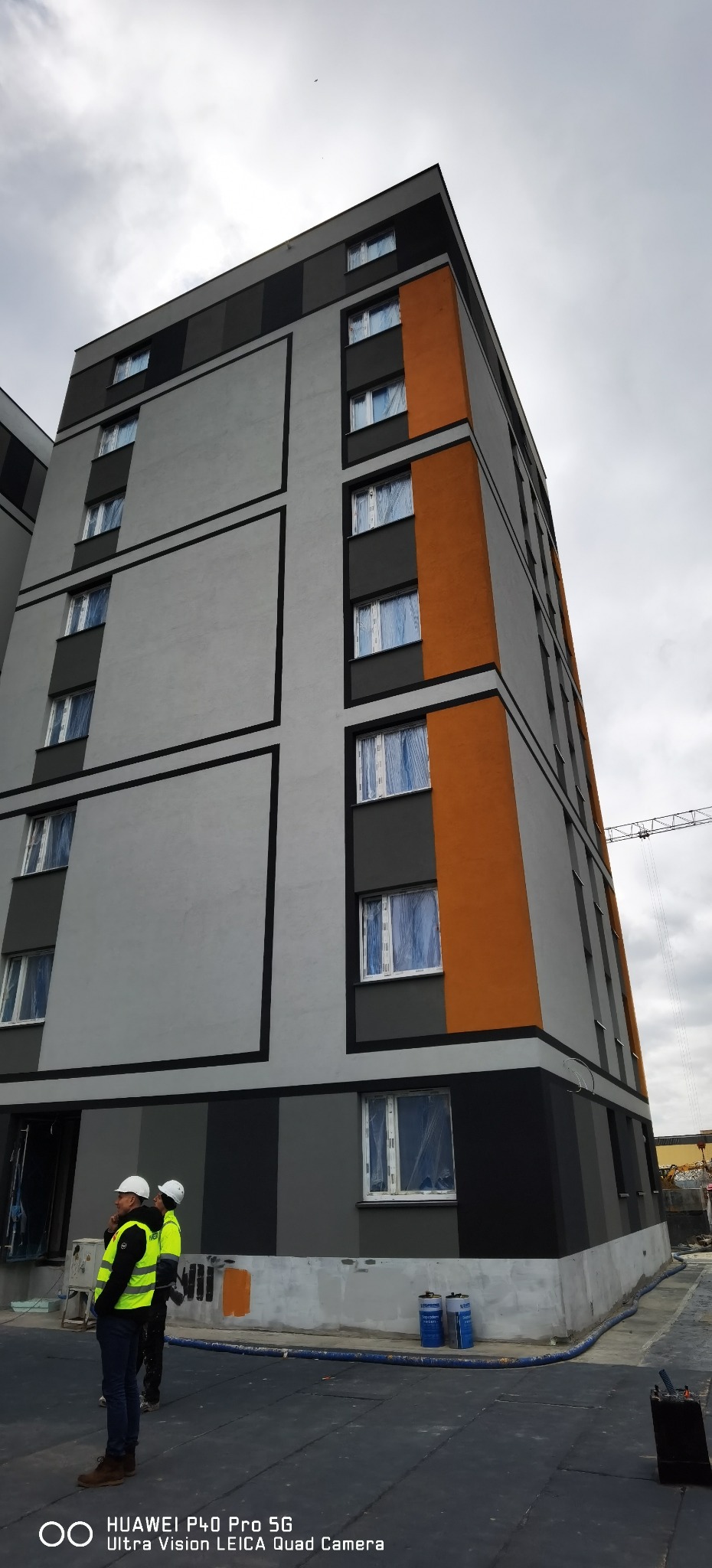 Wielopiętrowy budynek po renowacji elewacji w odcieniach szarości, czerni i pomarańczy. Okna zabezpieczone folią. Dwóch pracowników w kaskach i kamizelkach odblaskowych.