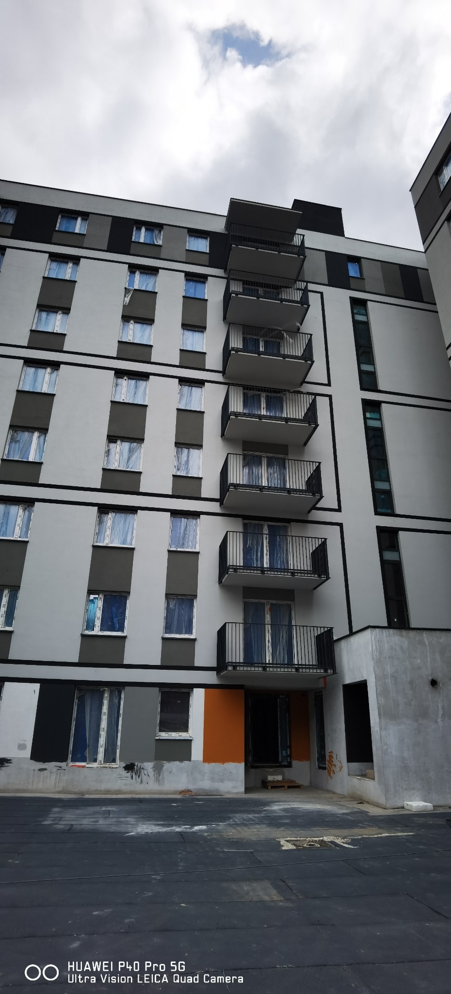 Wielopiętrowy budynek z balkonami w trakcie prac wykończeniowych. Okna zabezpieczone folią, widoczne fragmenty elewacji w różnych kolorach. Ujęcie z dołu.