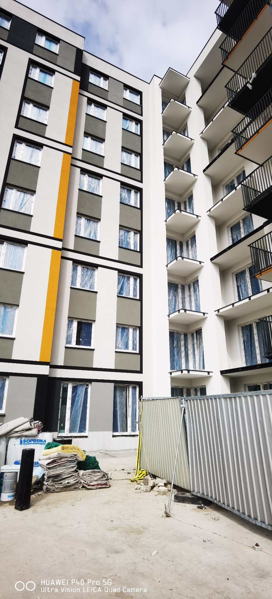 Wielopiętrowy budynek po remoncie elewacji z pomalowanymi balkonami i oknami zabezpieczonymi folią. Na dole widoczne materiały budowlane i tymczasowe ogrodzenie.