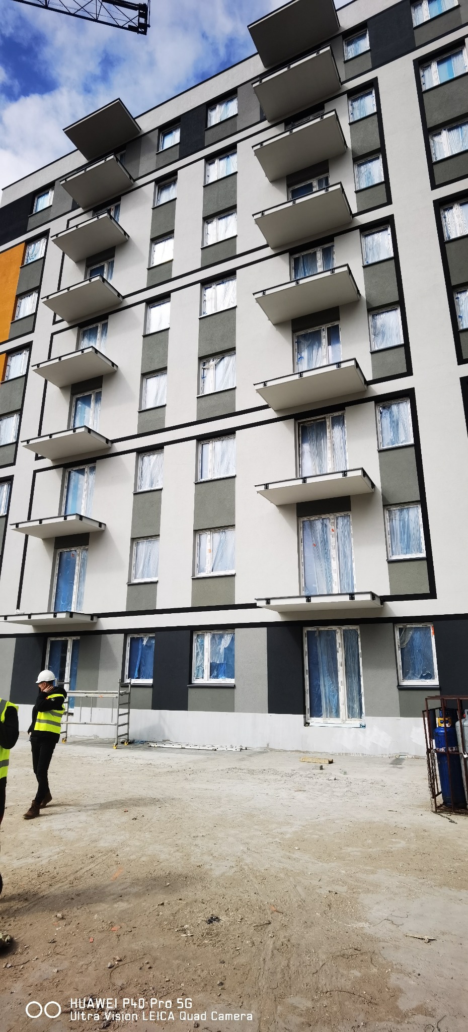 Wielopiętrowy budynek z balkonami w trakcie prac wykończeniowych. Okna zabezpieczone folią, widoczni pracownicy w odblaskowych kamizelkach i kaskach.