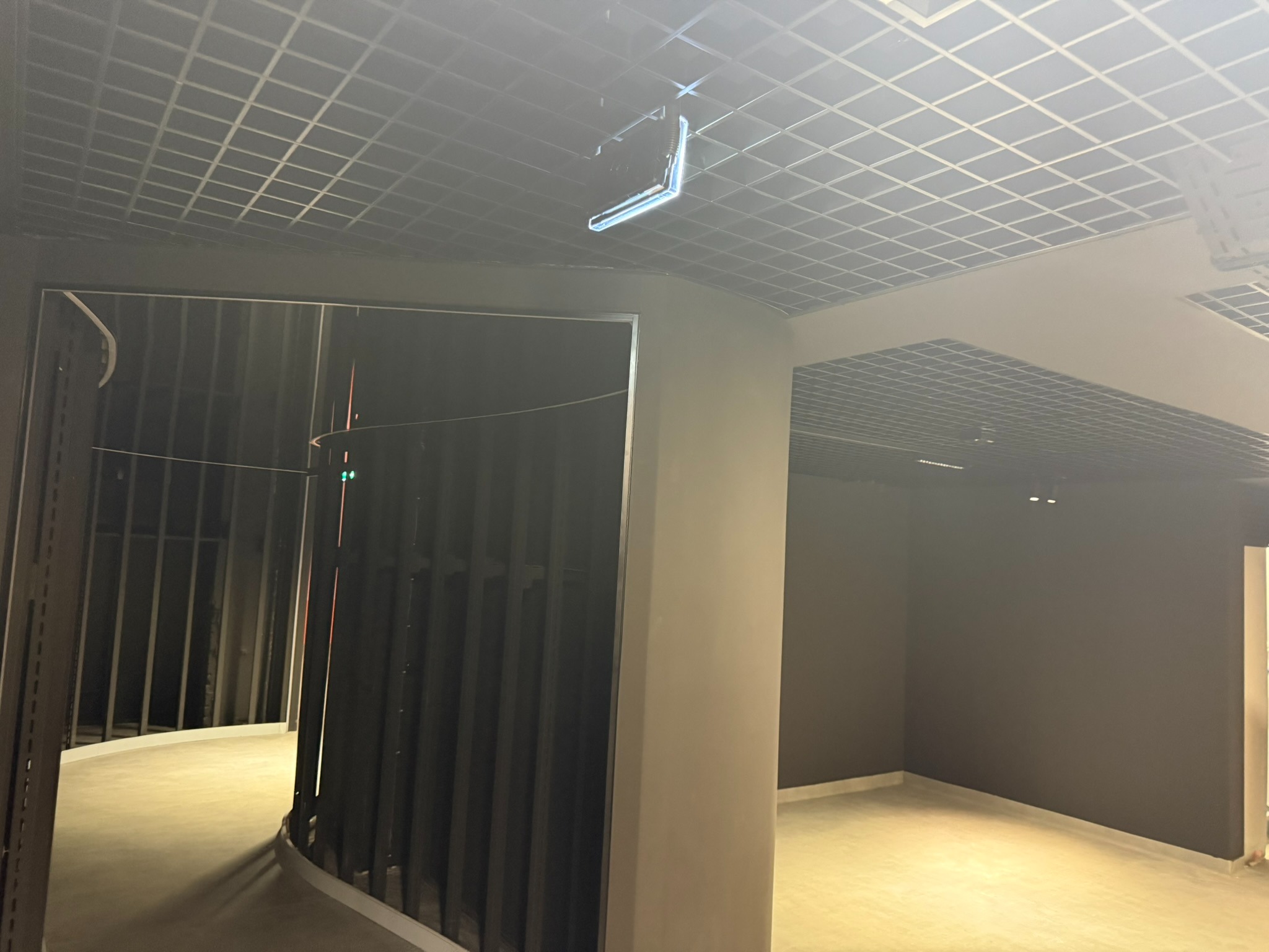 Wnętrze w trakcie wykańczania: sufit rastrowy, ściany w ciemnym kolorze, widoczne elementy konstrukcyjne. Oświetlenie sufitowe. Minimalistyczny design w surowym stanie.