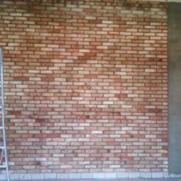 Arbud Arkadiusz Kufera - Ściana z cegły klinkierowej w trakcie montażu, częściowo otynkowana, z widoczną aluminiową drabiną i fragmentem podłogi.