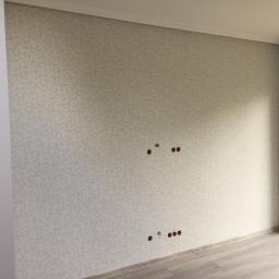 Arbud Arkadiusz Kufera - Nowo wykończone wnętrze pokoju z wzorzystą tapetą w odcieniach szarości, widoczne puszki elektryczne bez osprzętu i panele podłogowe imitujące drewno.