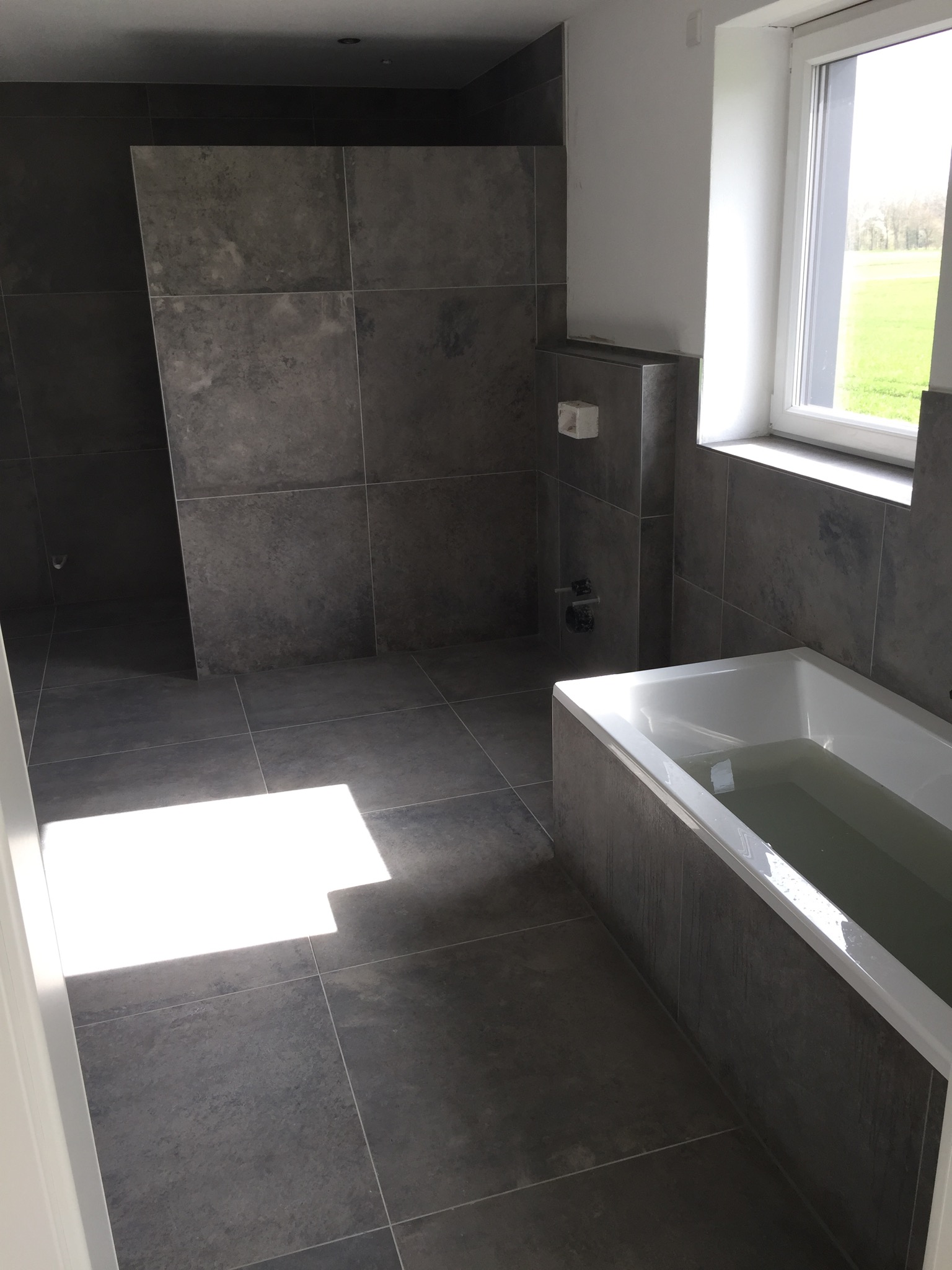 Nowoczesna łazienka z szarymi płytkami wielkoformatowymi na ścianach i podłodze, biała wanna wypełniona wodą, okno z widokiem na zieleń za oknem.