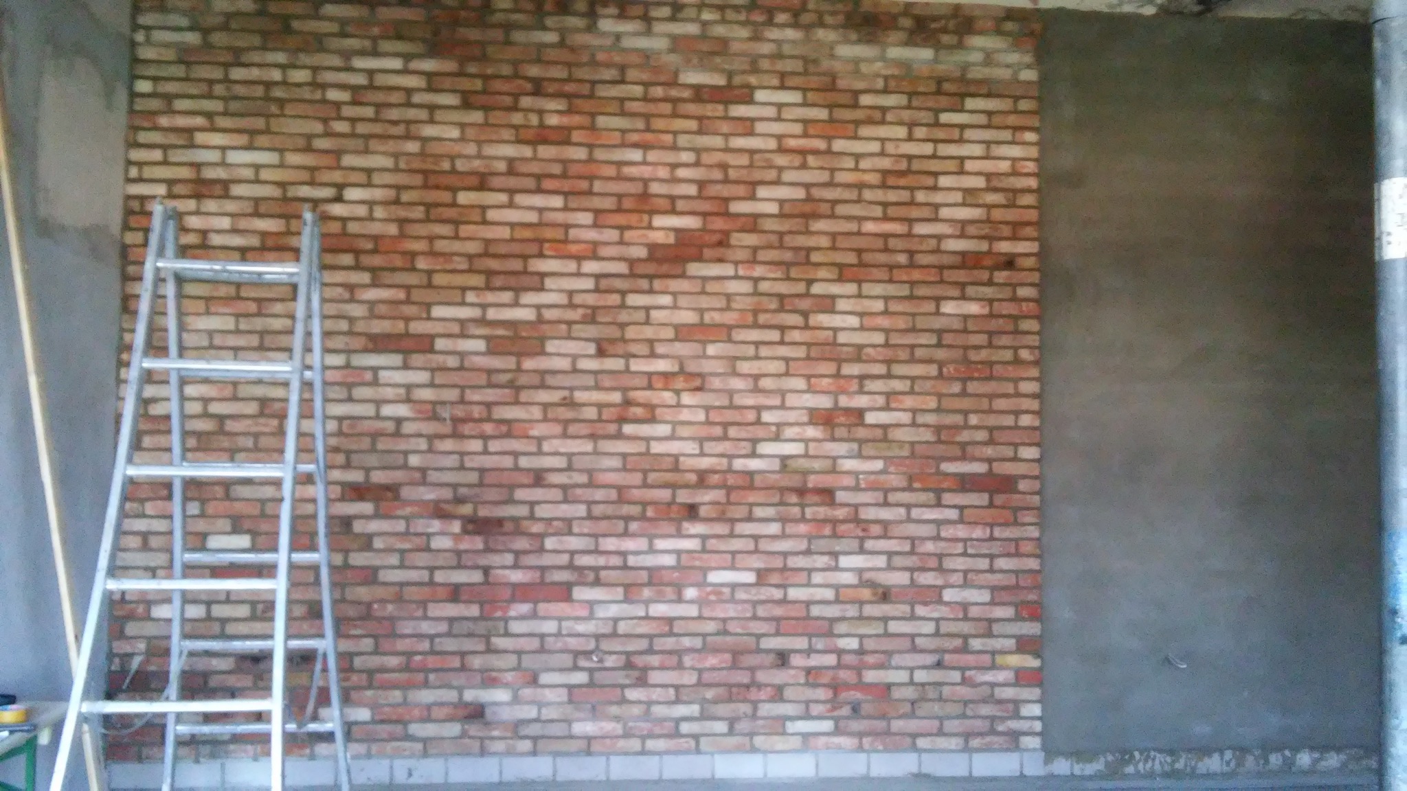 Ściana z cegły klinkierowej w trakcie montażu, częściowo otynkowana, z widoczną aluminiową drabiną i fragmentem podłogi.