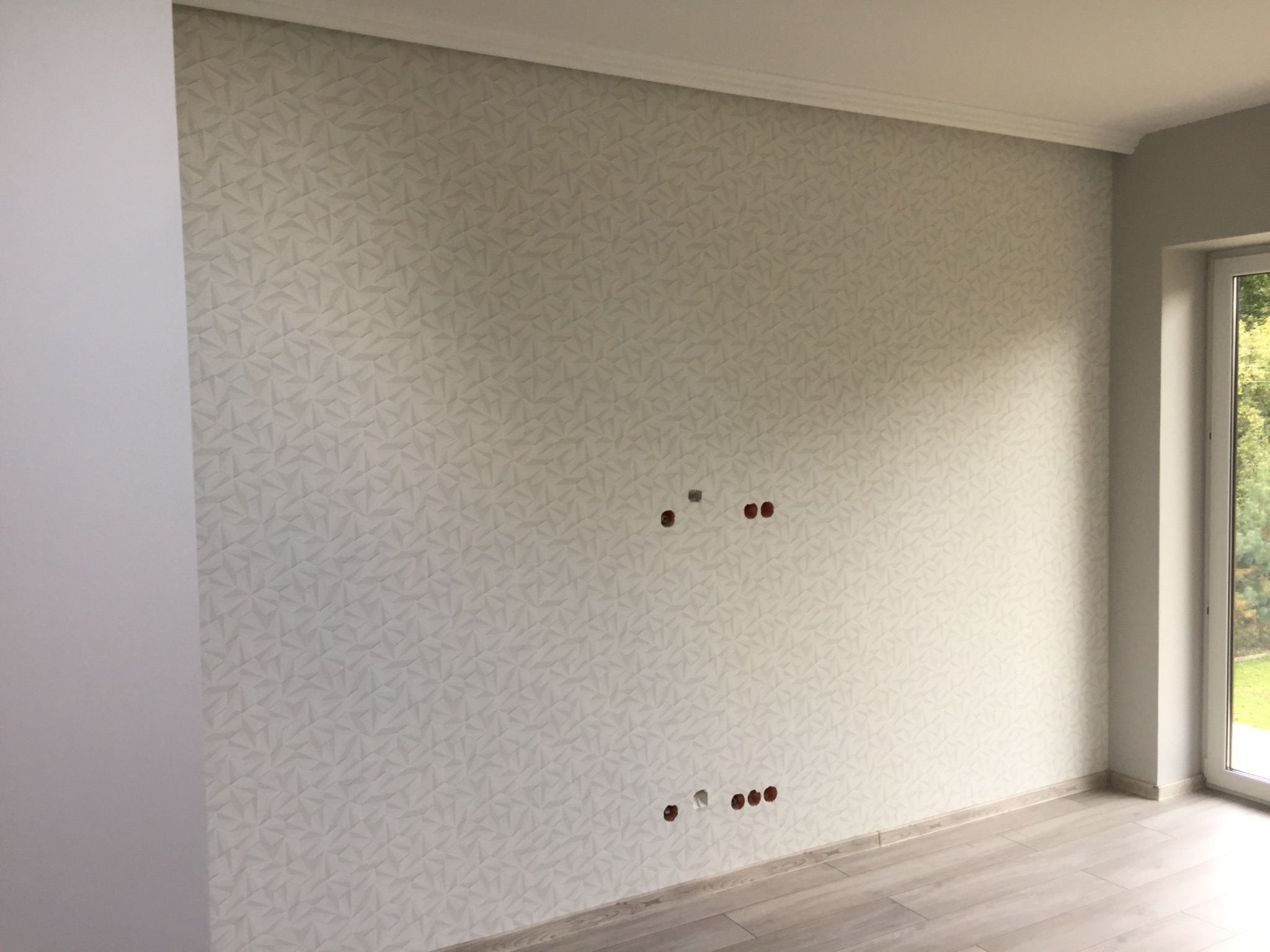 Nowo wykończone wnętrze pokoju z wzorzystą tapetą w odcieniach szarości, widoczne puszki elektryczne bez osprzętu i panele podłogowe imitujące drewno.