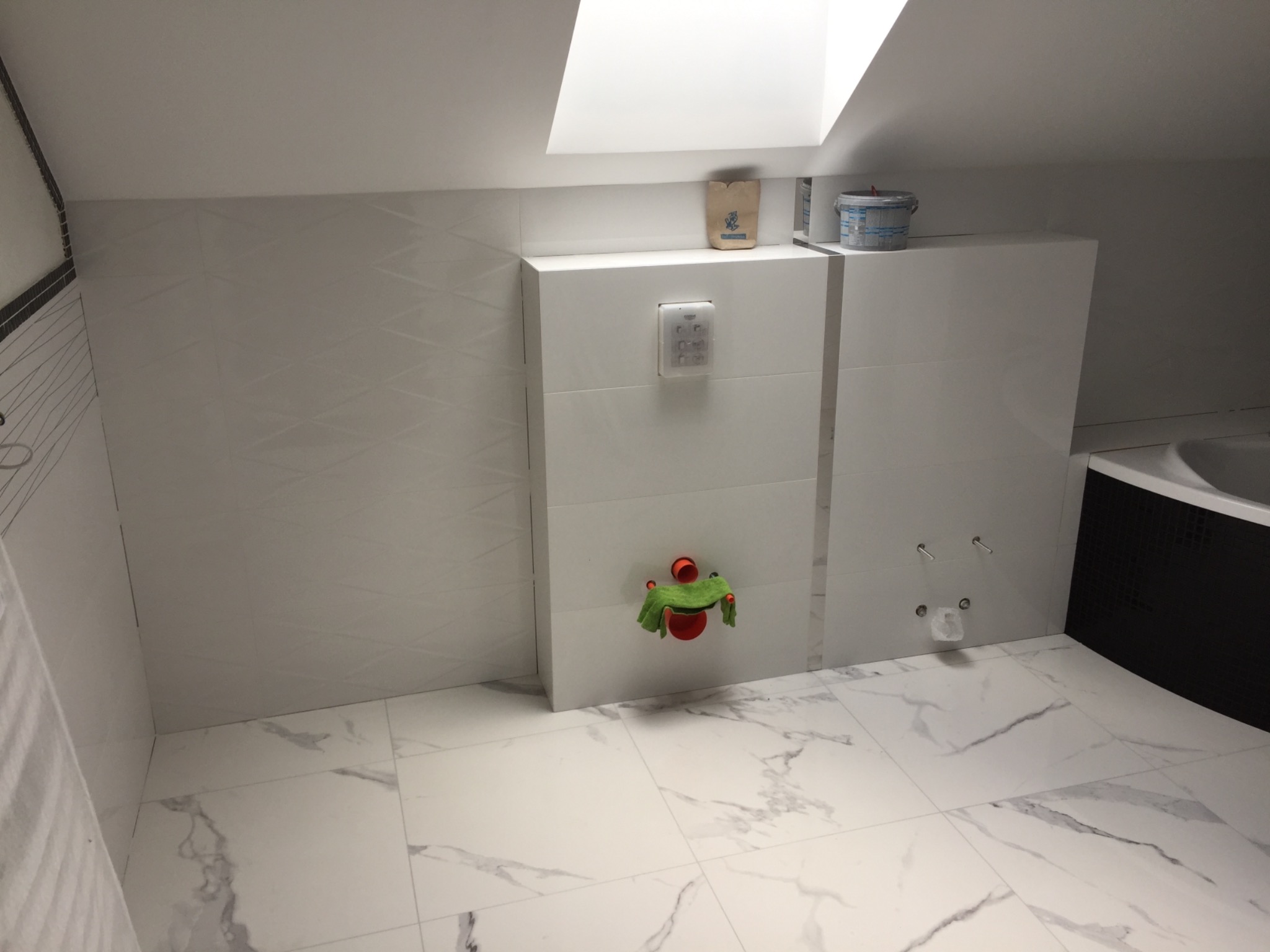 Wykończona łazienka na poddaszu z białymi płytkami imitującymi marmur na podłodze i ścianach, widoczna zabudowa stelaża WC z przyciskiem spłukującym, workiem z klejem i wiadrem z farbą na górze...