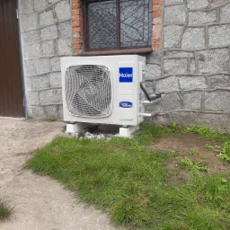 Biała pompa ciepła Haier zamontowana na zewnątrz budynku z kamienną elewacją, widoczny fragment trawnika i okno z ozdobną kratą.